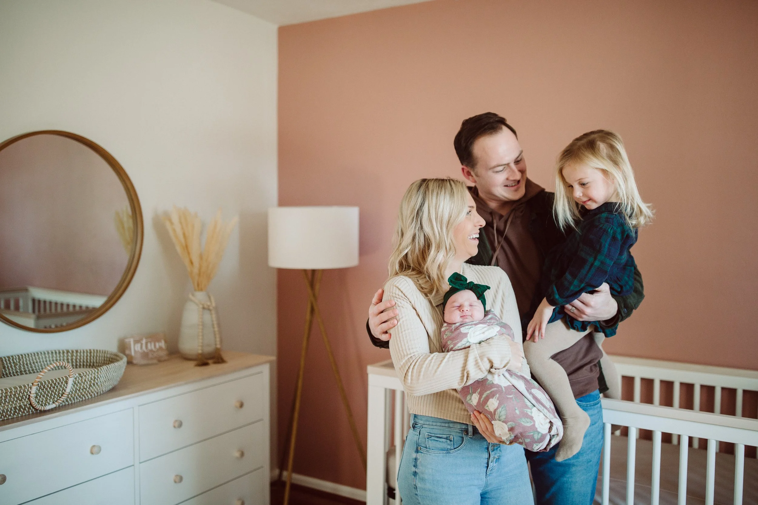A smiling family of four stands in a nursery. The mother holds a newborn girl wrapped in a floral blanket, and the father lifts a young girl, all smiling at each other. The nursery has a pink accent wall, a white dresser, a round mirror, a lamp, and
