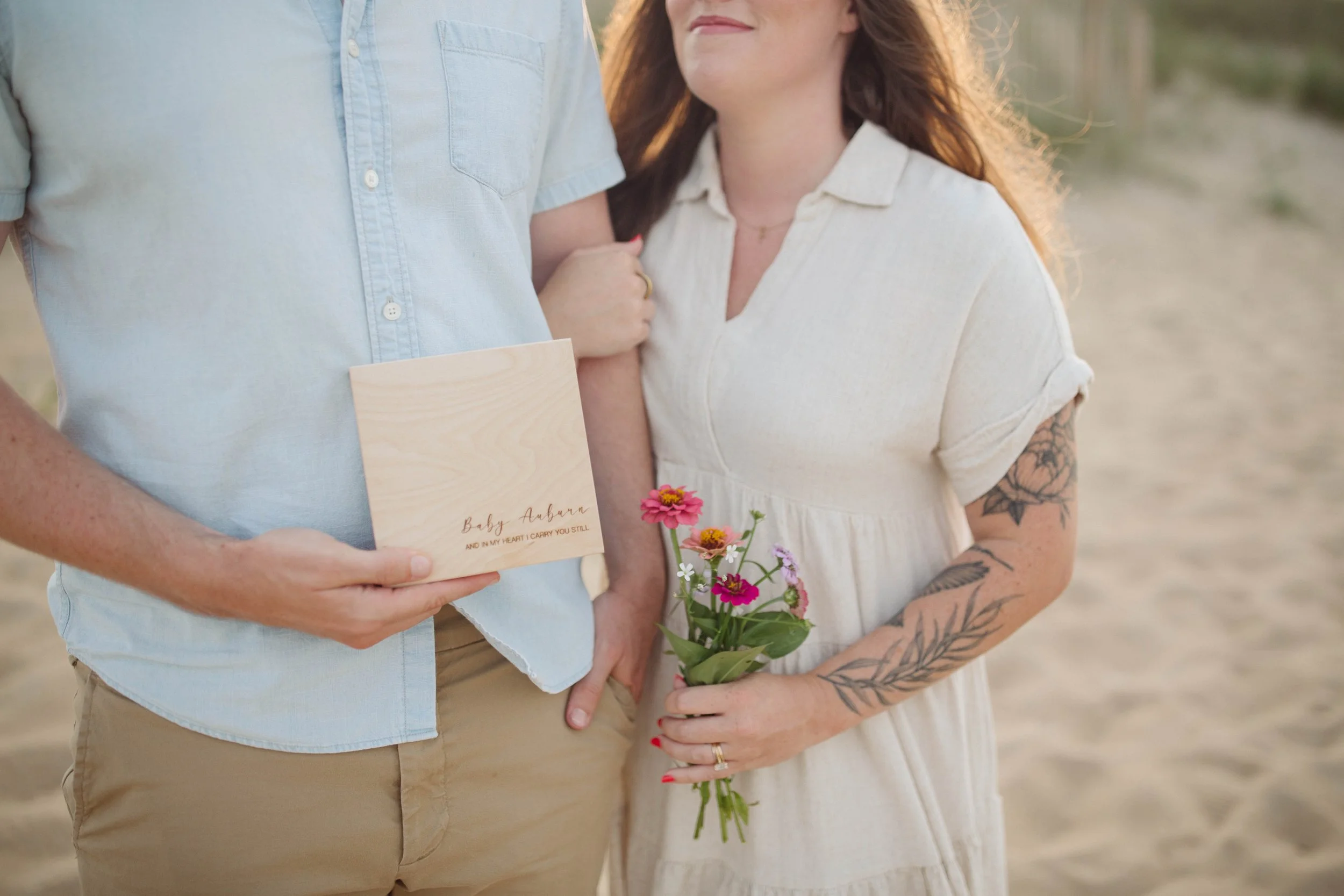 A couple stands on the beach during sunset. The man holds a wooden card with the writing, "Baby Auburn" and an additional message. The woman holds a small bouquet of pink and white flowers, with visible tattoos on her left arm.