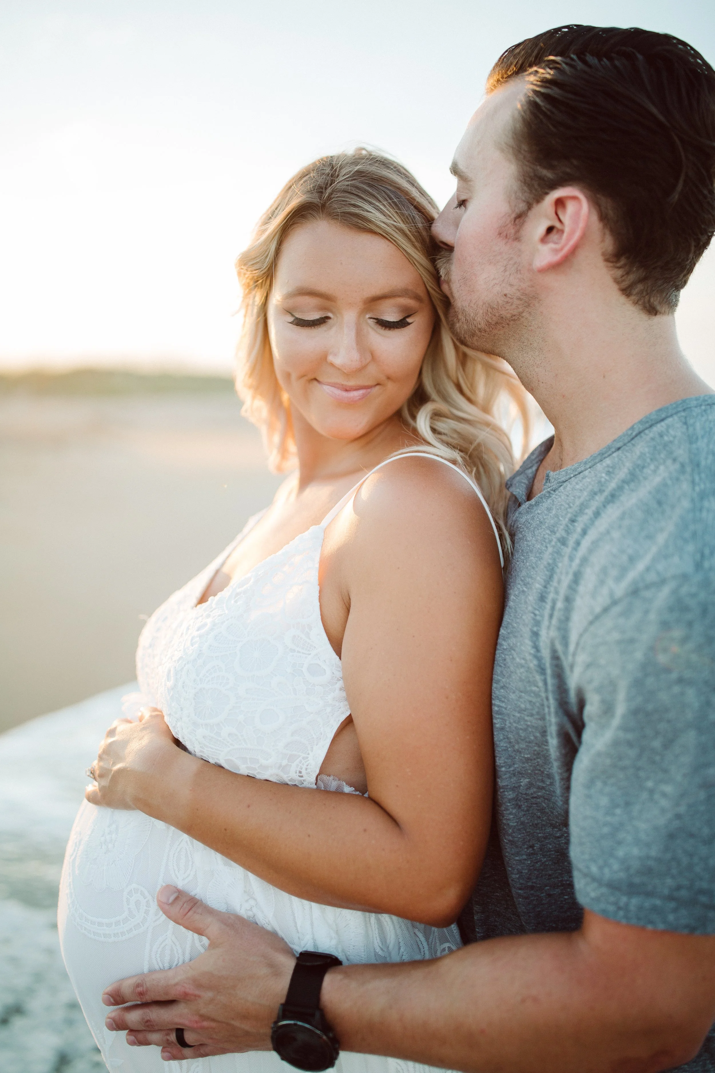 A pregnant woman in a white dress standing on the beach with a man in a gray shirt kissing her forehead, both with eyes closed, during sunset.