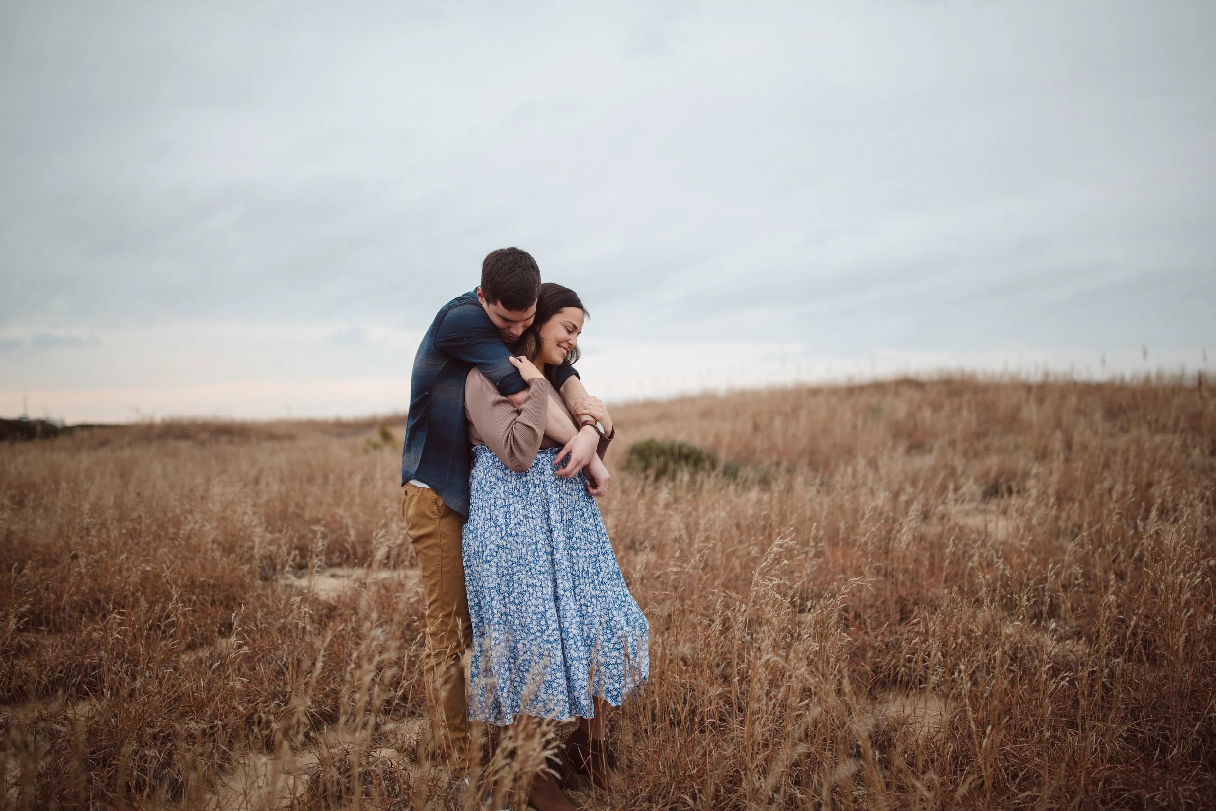 A couple embraces in a field of tall, dry grass under a cloudy sky.