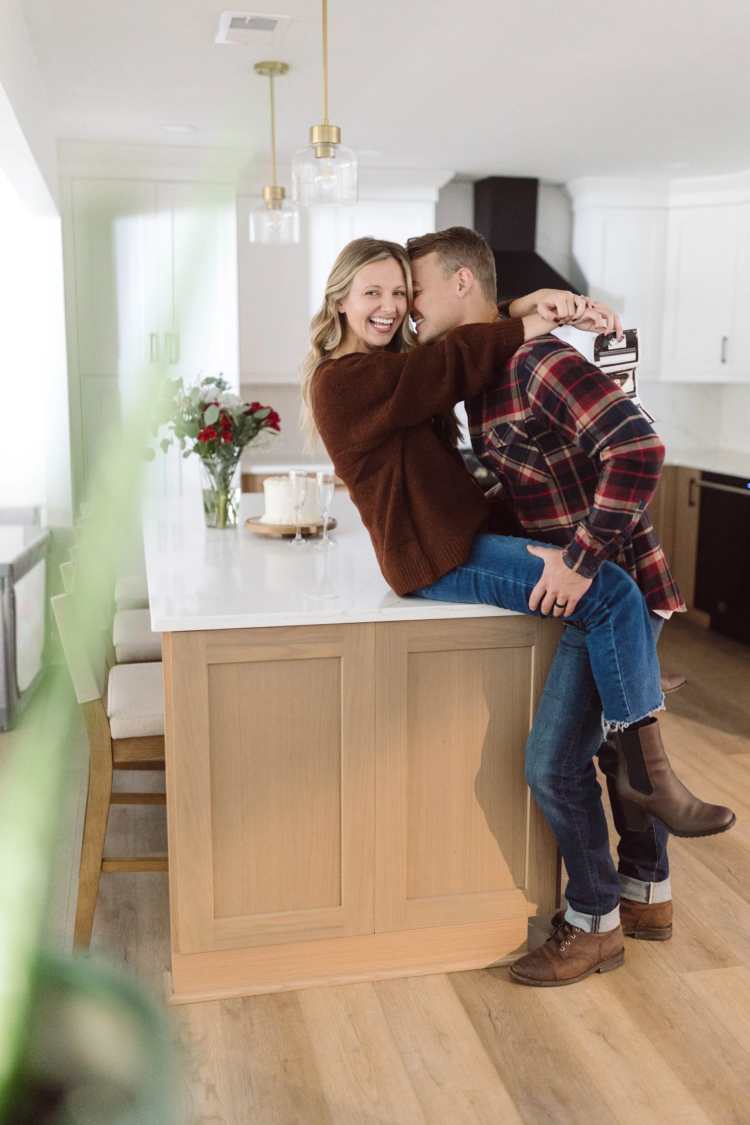 A smiling woman and man embrace and laugh in a kitchen. The woman is sitting on a kitchen island, and the man is leaning in to her.