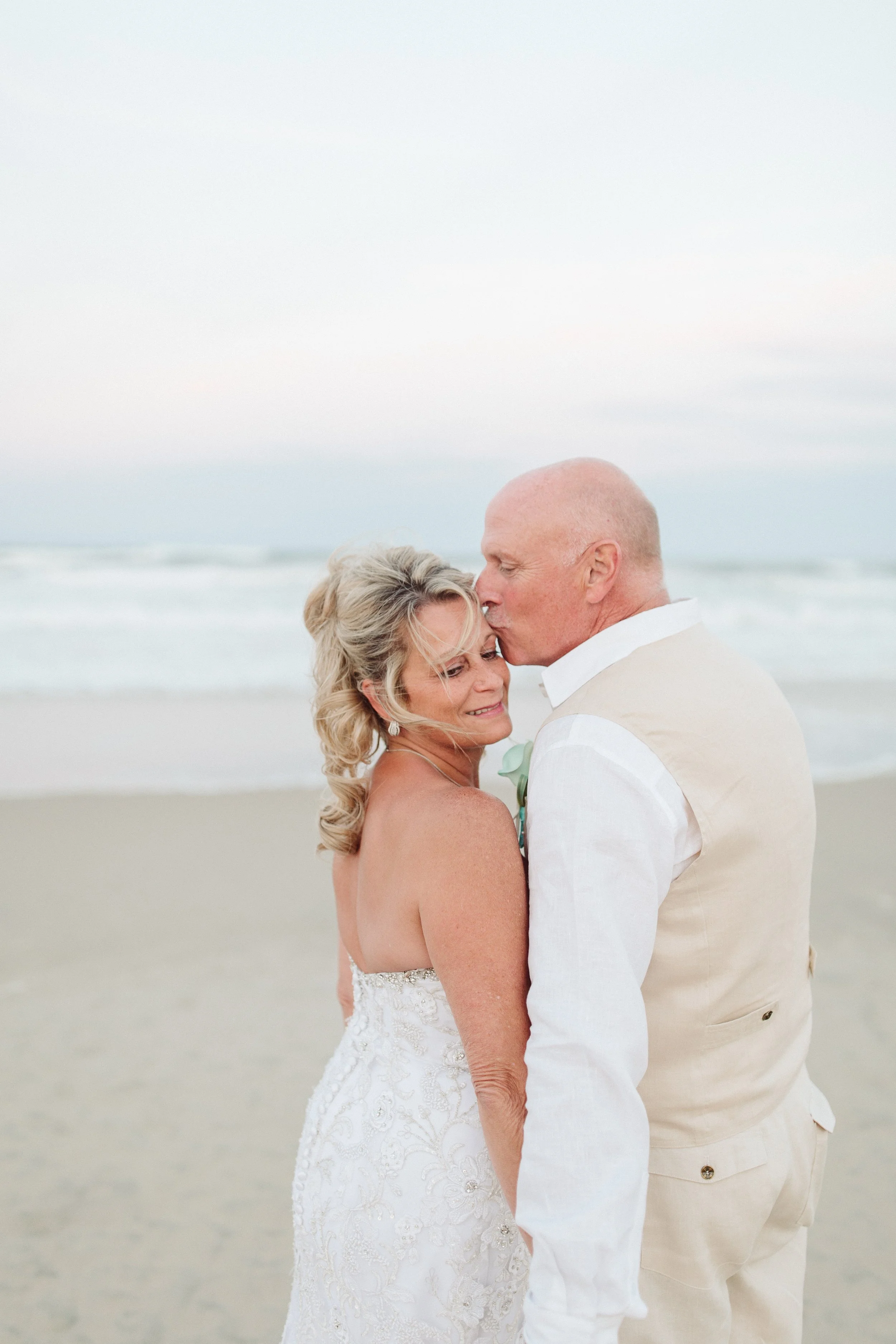 A bride and groom embrace on the beach, the groom kissing the bride on her forehead during sunset or early evening.