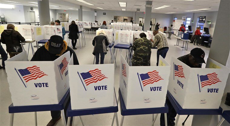 People casting votes at a polling station with voting booths decorated with American flags and the word 'VOTE'.