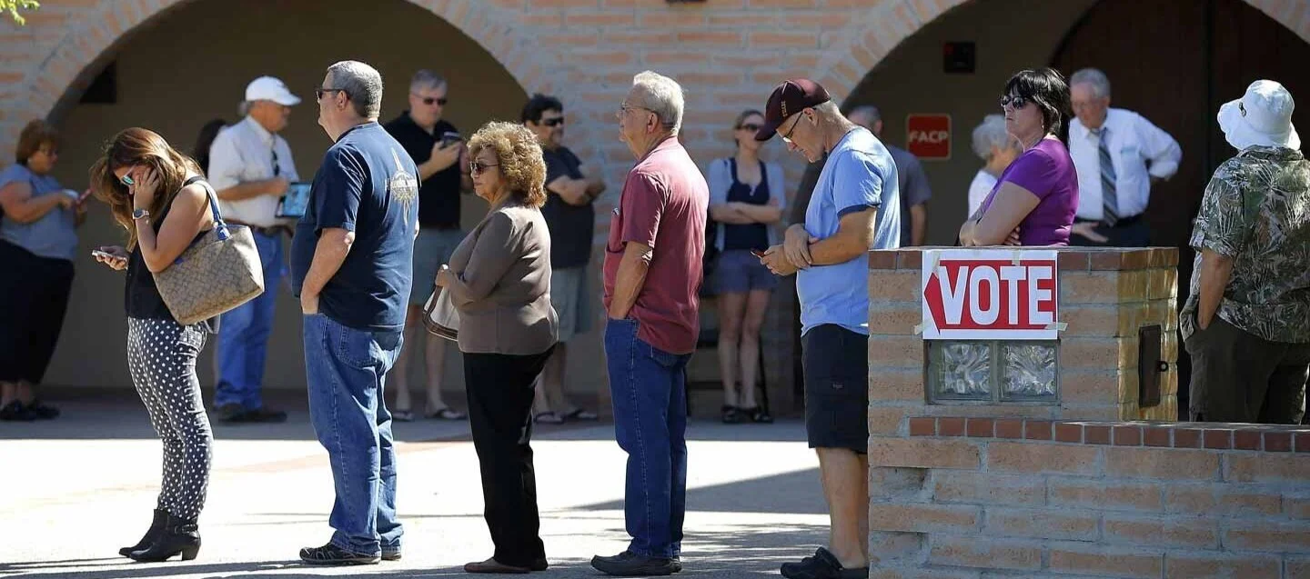 People standing in a line outside a voting station on a sunny day, with a brick wall and a 'VOTE' sign in the background.