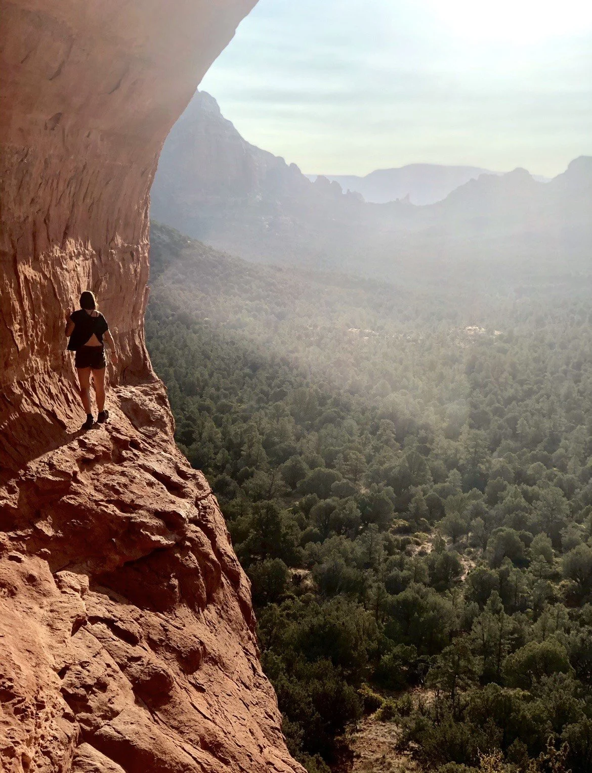 the wild west and the frontier of intuitive wisdom,  person climbing along a narrow ledge on a red rock cliff overlooking a green forest and distant mountains.