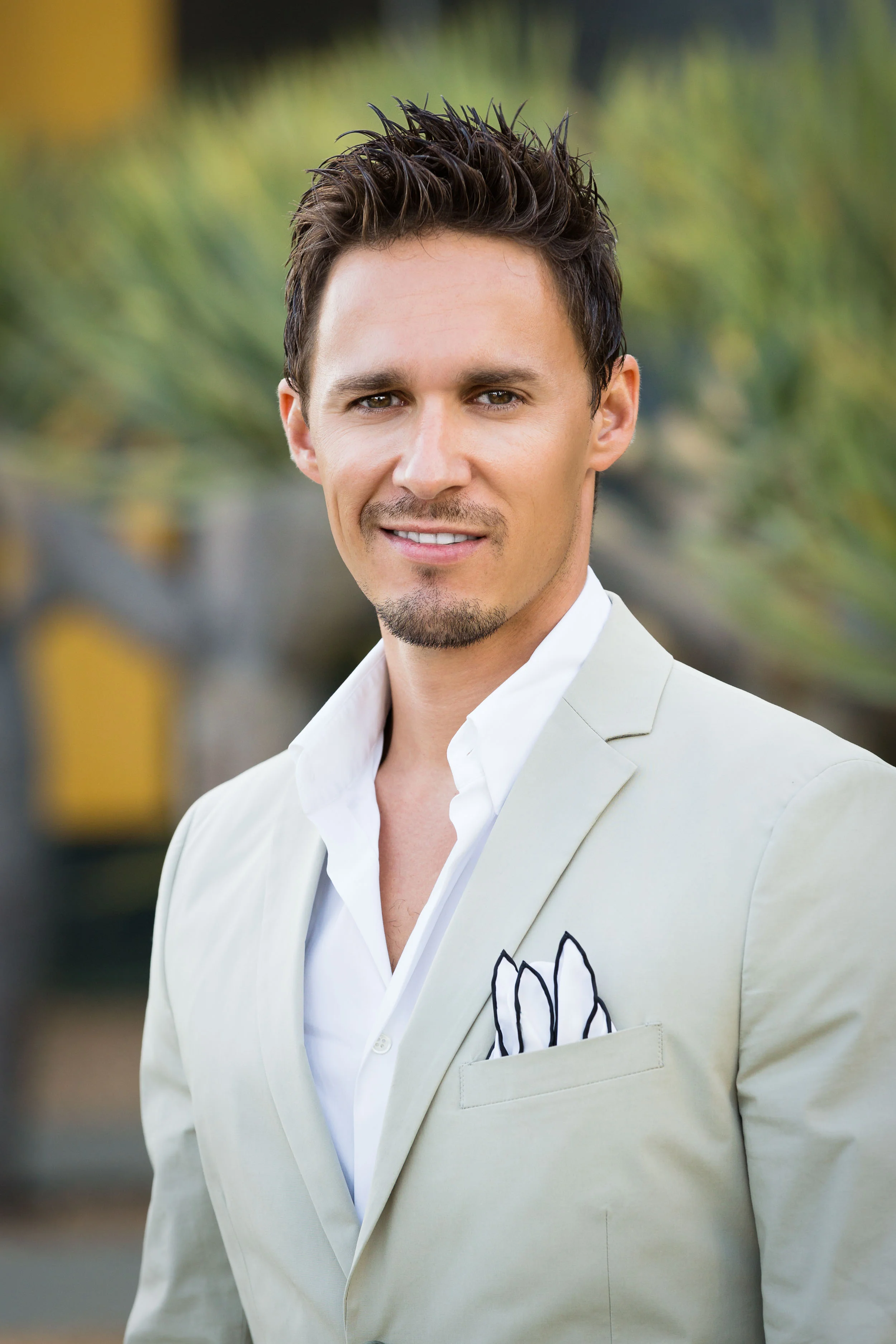 Man in a light beige suit with a white shirt, smiling outdoors with a blurred green background, in a professional environmental portrait in Los Angeles.