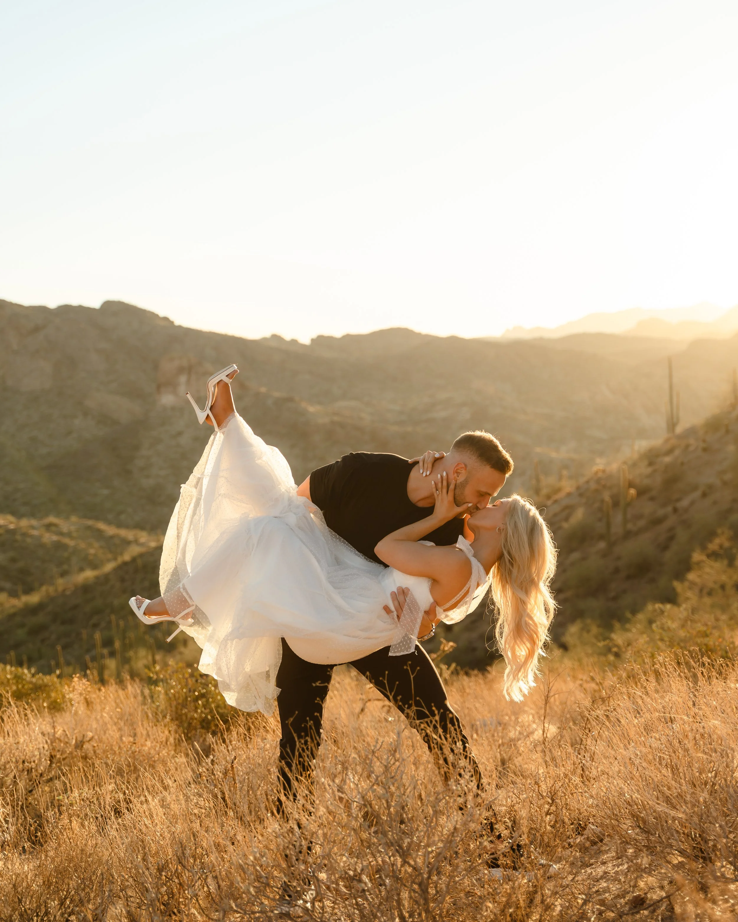 A man in black clothing and a woman in a white dress with high heels are outdoors at sunset in a desert landscape, with the man lifting the woman in his arms and they are about to kiss.
