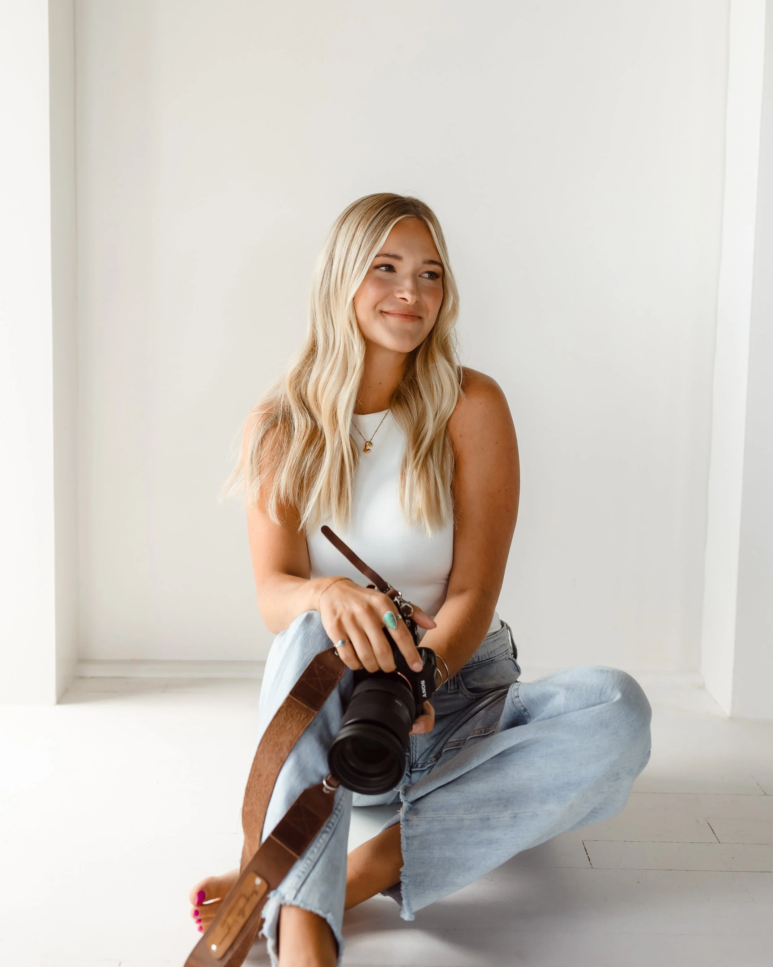 A young woman with long blonde hair, wearing a white top and light jeans, sitting cross-legged on the floor with a camera in her hands, smiling and looking slightly to her left, in a minimalist white room.