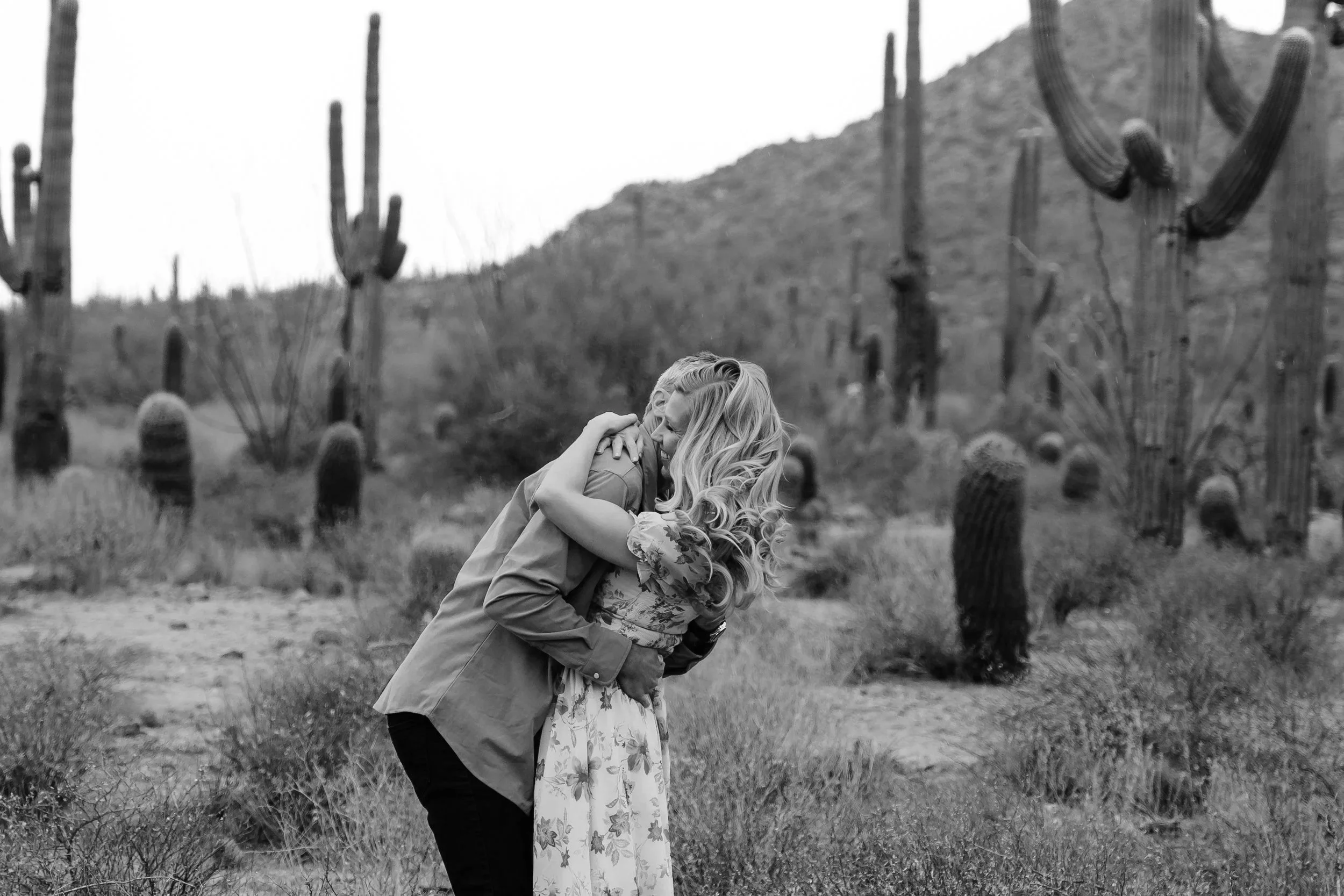 A couple embracing and smiling in a desert landscape with tall saguaro cacti and dry bushes.