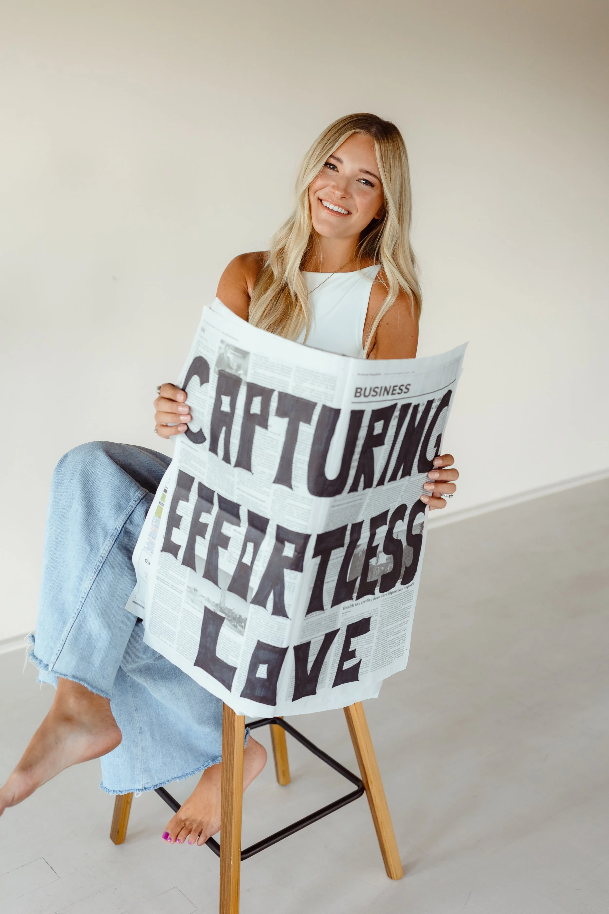 A woman sitting on a stool, smiling, holding a newspaper with large text about cheering, happiness, and love.