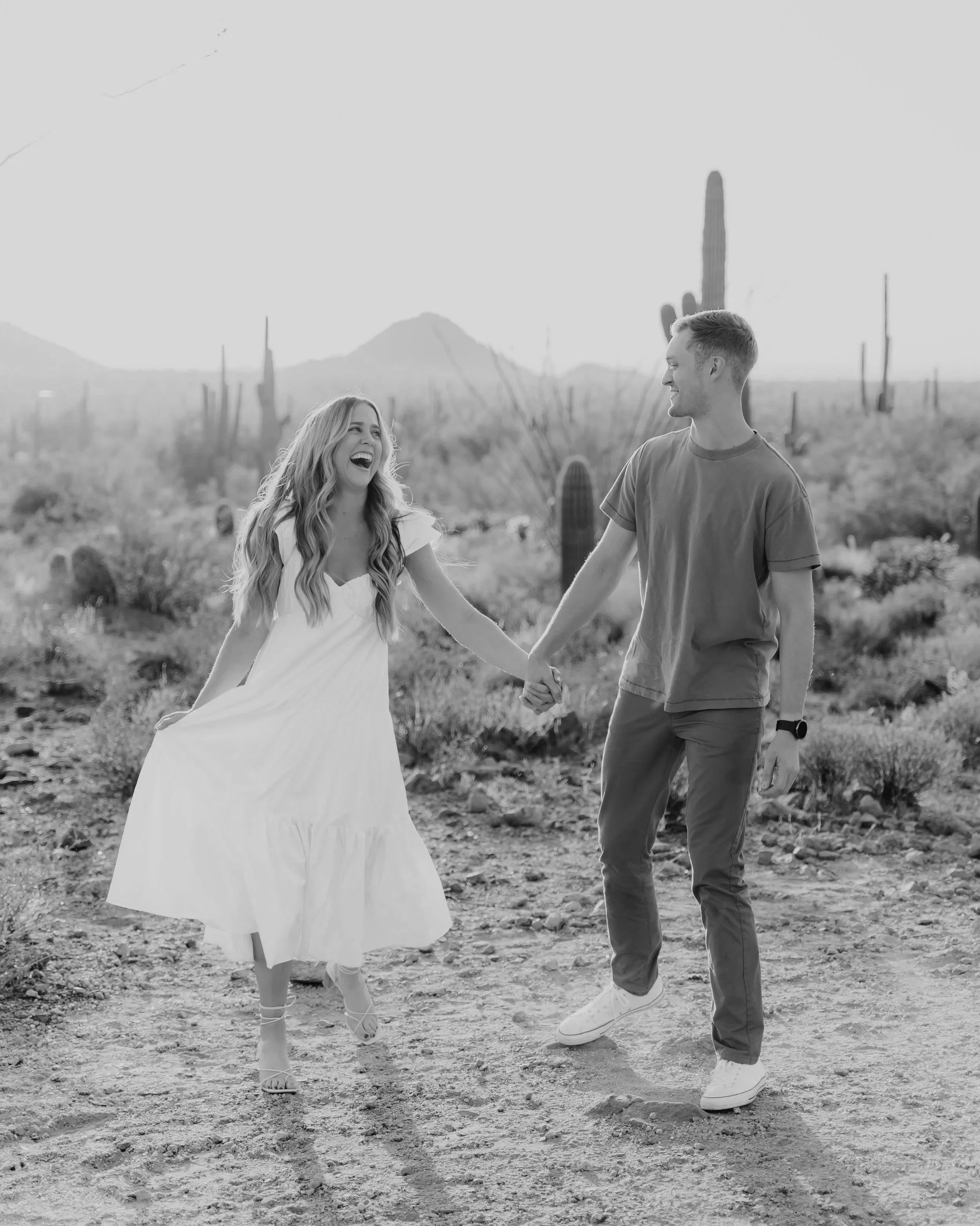 A smiling woman in a white dress and a man in a brown t-shirt holding hands and dancing in a desert with cacti and mountains in the background during sunset.