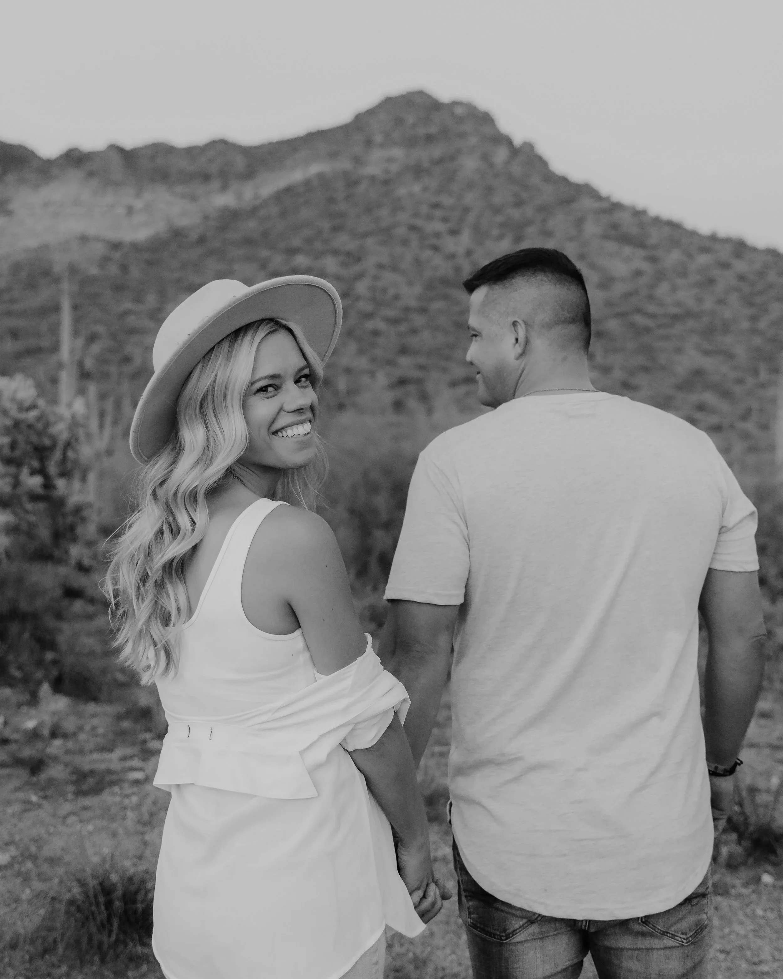 A smiling woman with long blonde hair wearing a wide-brimmed hat and white dress, holding hands with a man in a light-colored t-shirt in a desert landscape with mountains in the background.