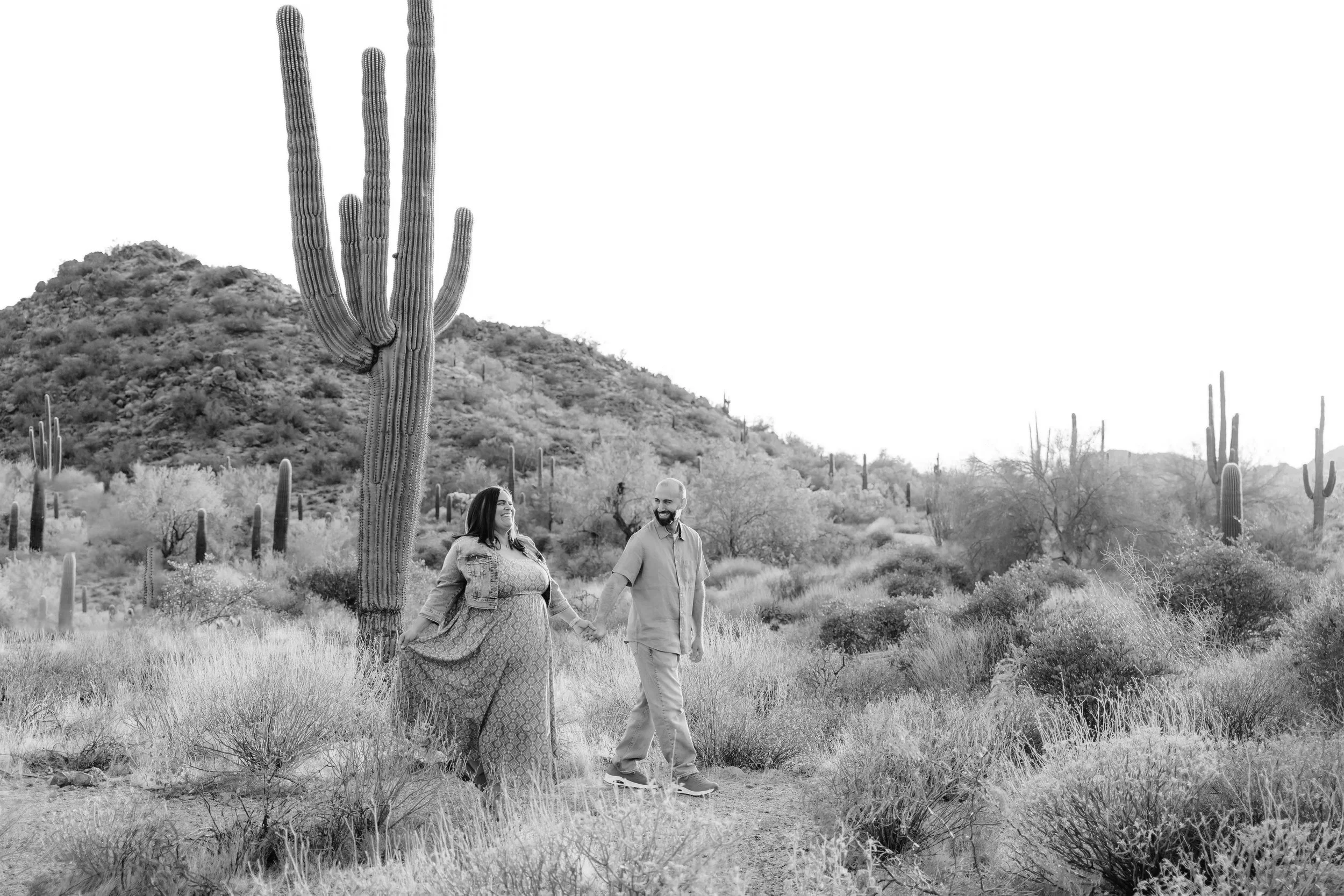 A couple holding hands and walking through a desert landscape with tall cacti and dry bushes at sunset.