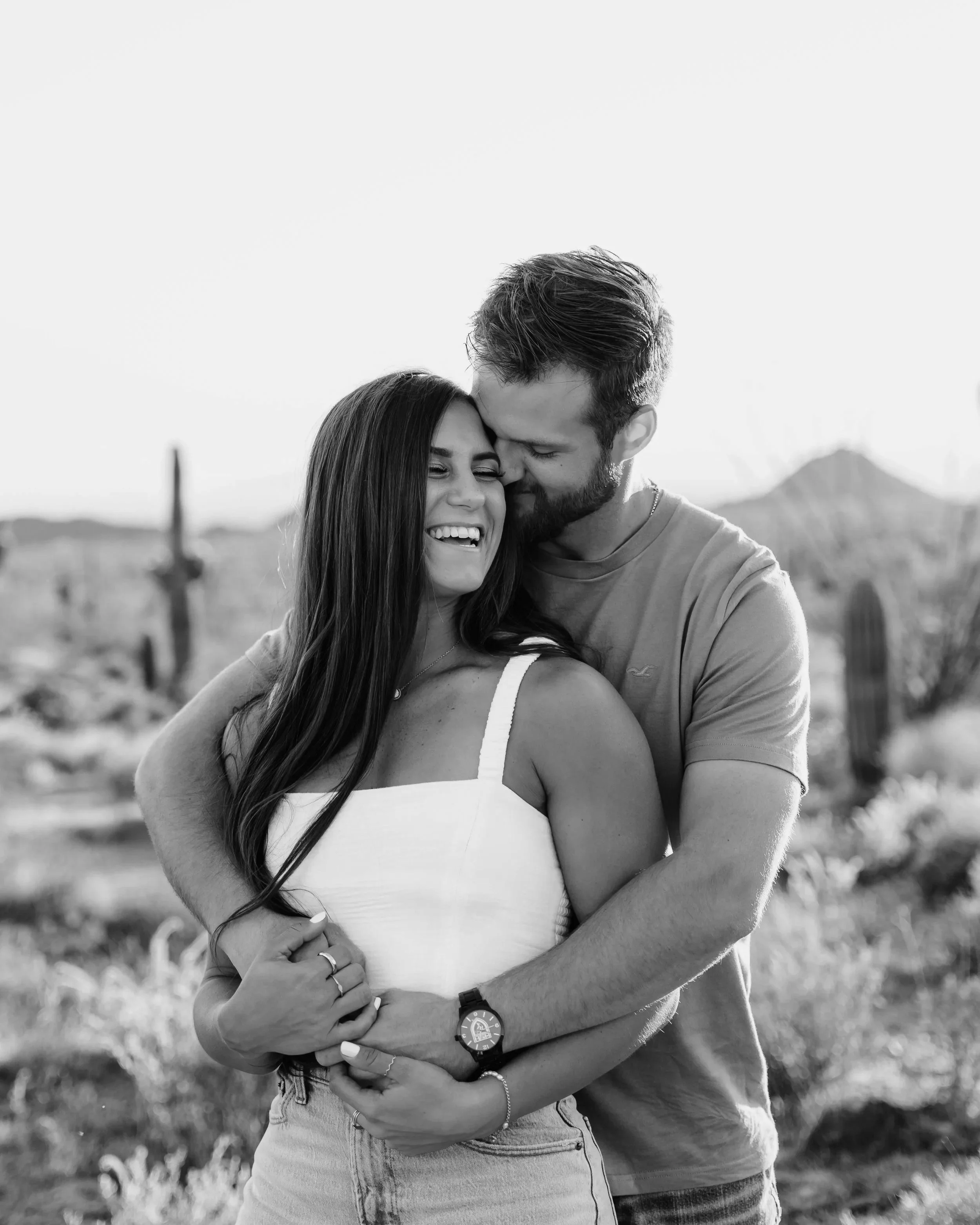 A couple hugging in a desert landscape with cacti, smiling happily, during sunset.
