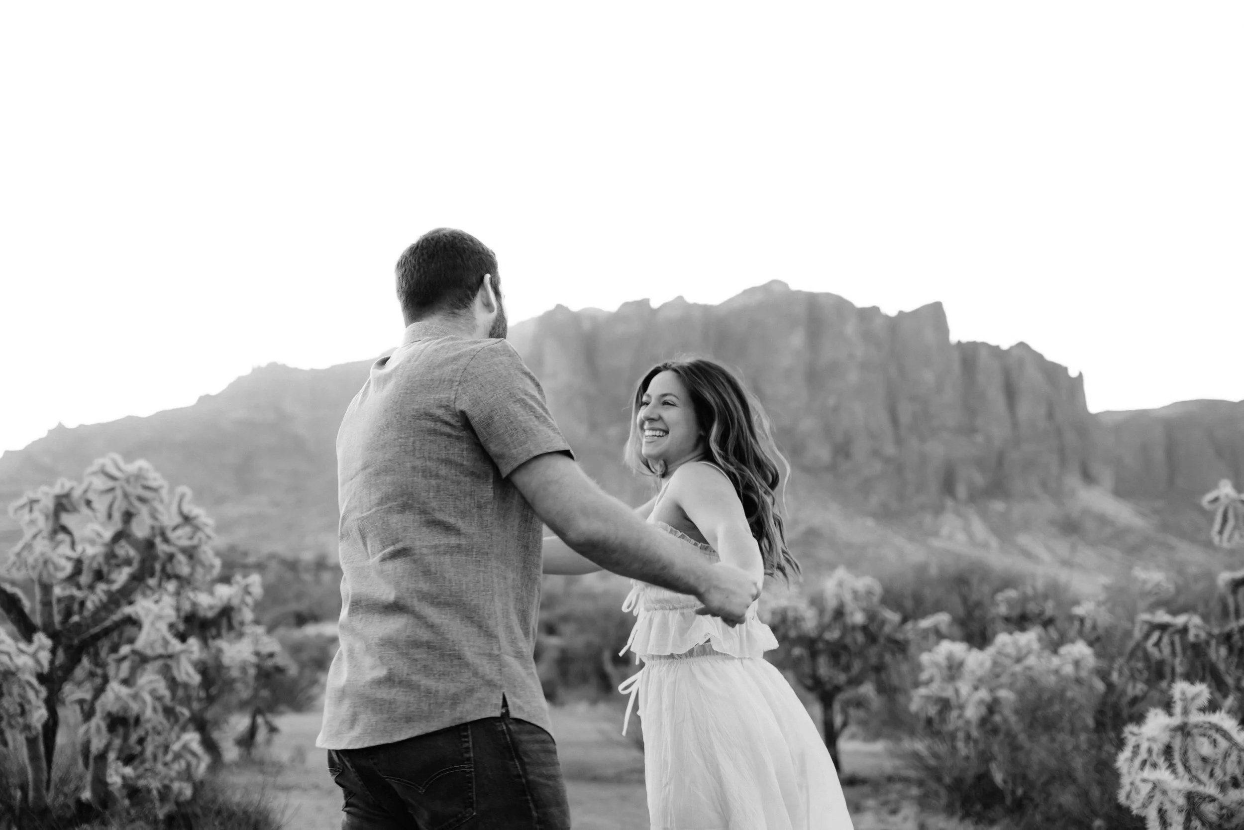 A couple dancing outdoors in a desert landscape with mountains in the background, the woman smiling and wearing a white dress, the man in a gray shirt.