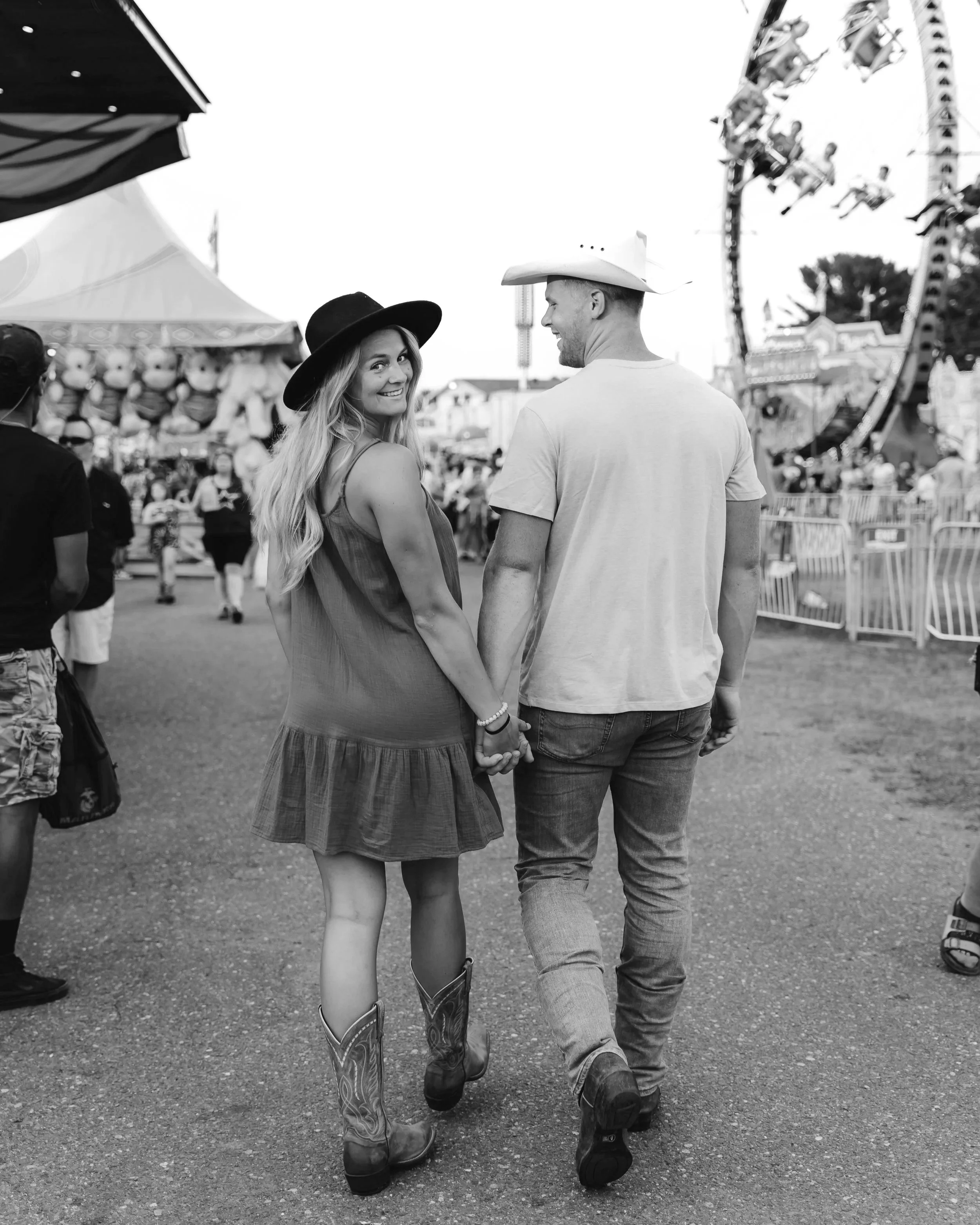 A smiling woman in a black hat and a reddish-brown dress holding hands with a man in a white cowboy hat and jeans at a fairground, with rides and booths in the background.