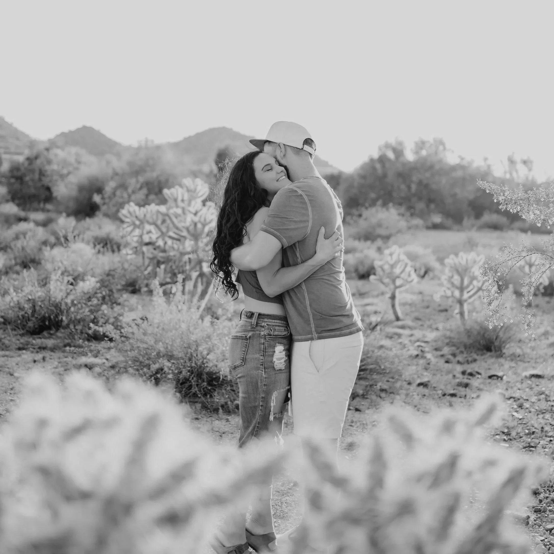 A couple hugging in a desert landscape with cacti and mountains in the background during sunset.