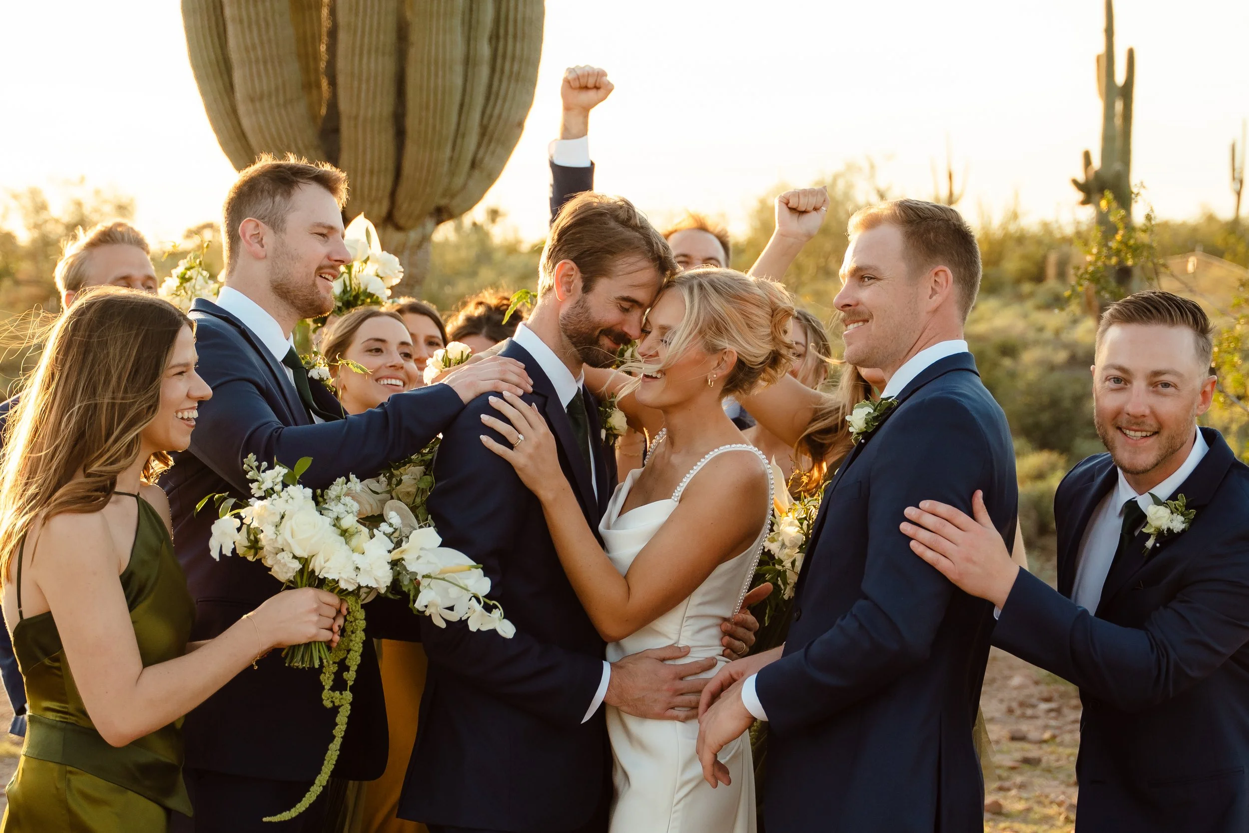 A wedding celebration outdoors with a bride and groom surrounded by friends, everyone smiling and hugging in front of a desert landscape with a big cactus during sunset.