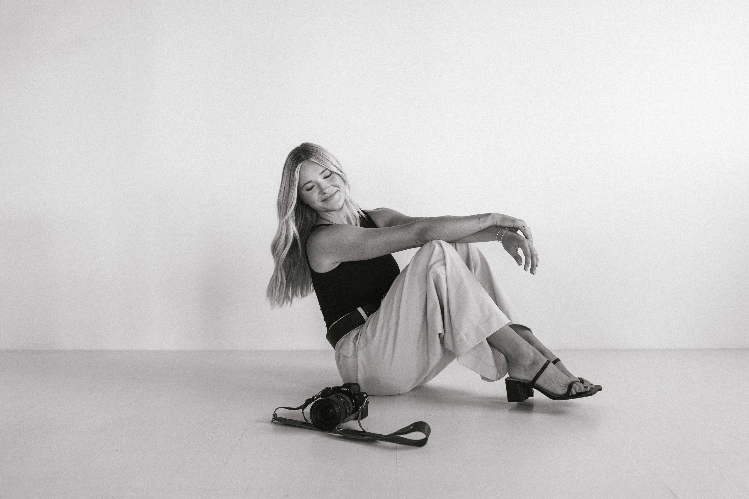A woman sitting on the floor with a camera next to her, smiling with eyes closed, in a minimal studio setting.