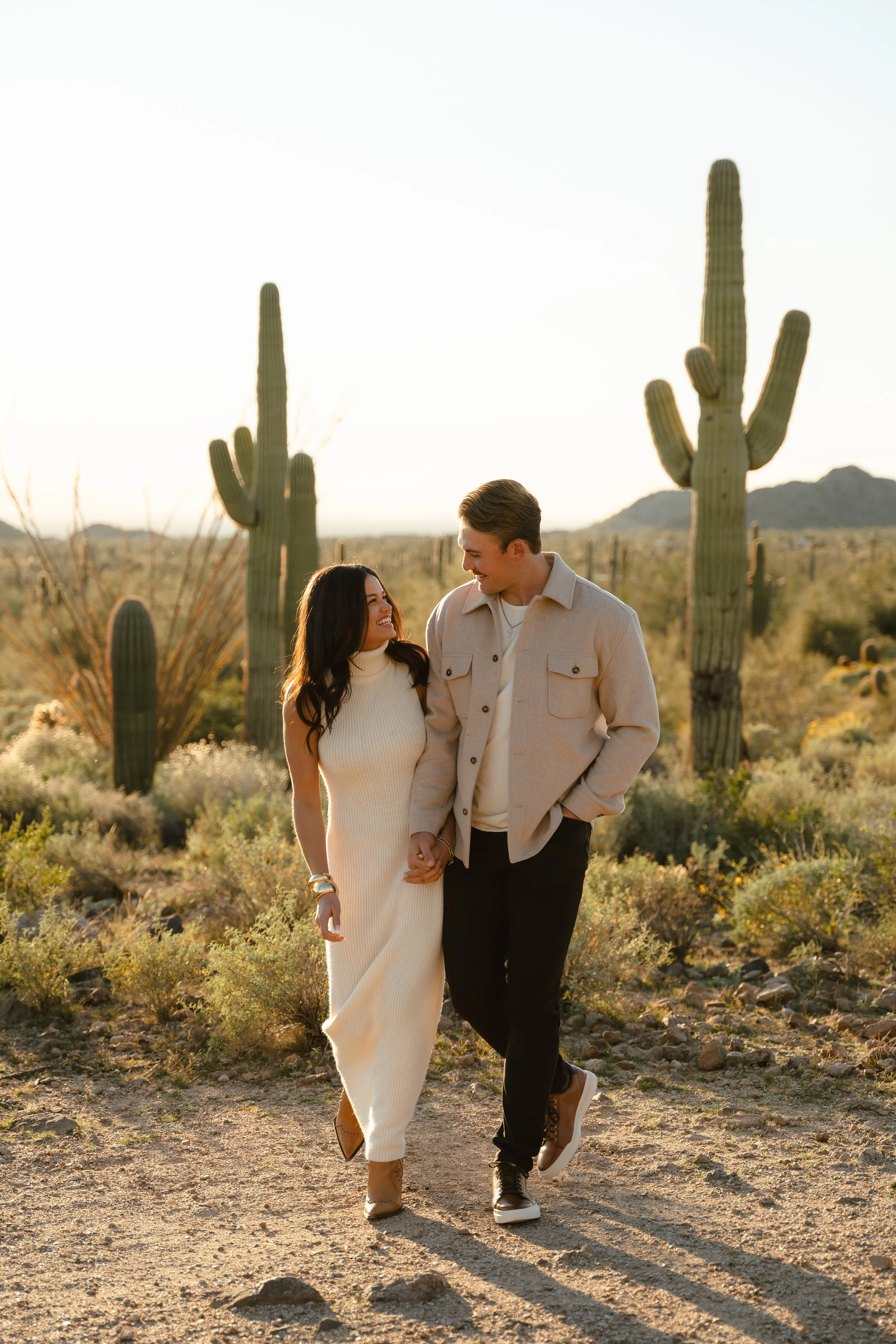 A couple walking hand in hand in a desert landscape with large saguaro cacti and mountains in the background, during sunset.