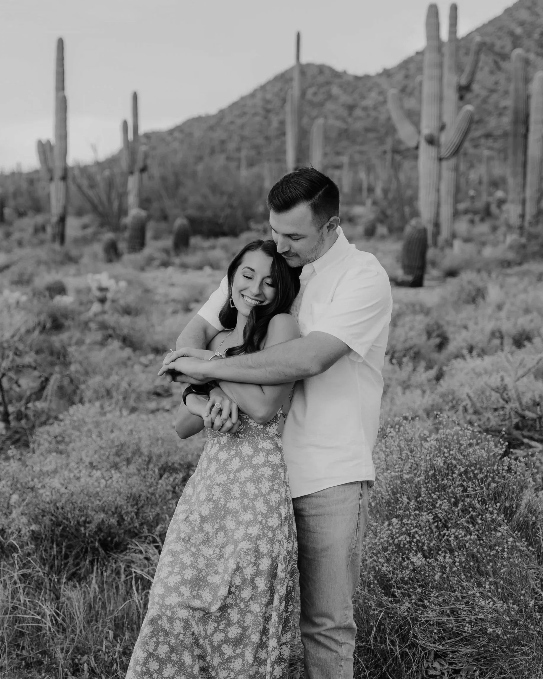A happy couple hugging in a desert landscape with cacti and mountains in the background.