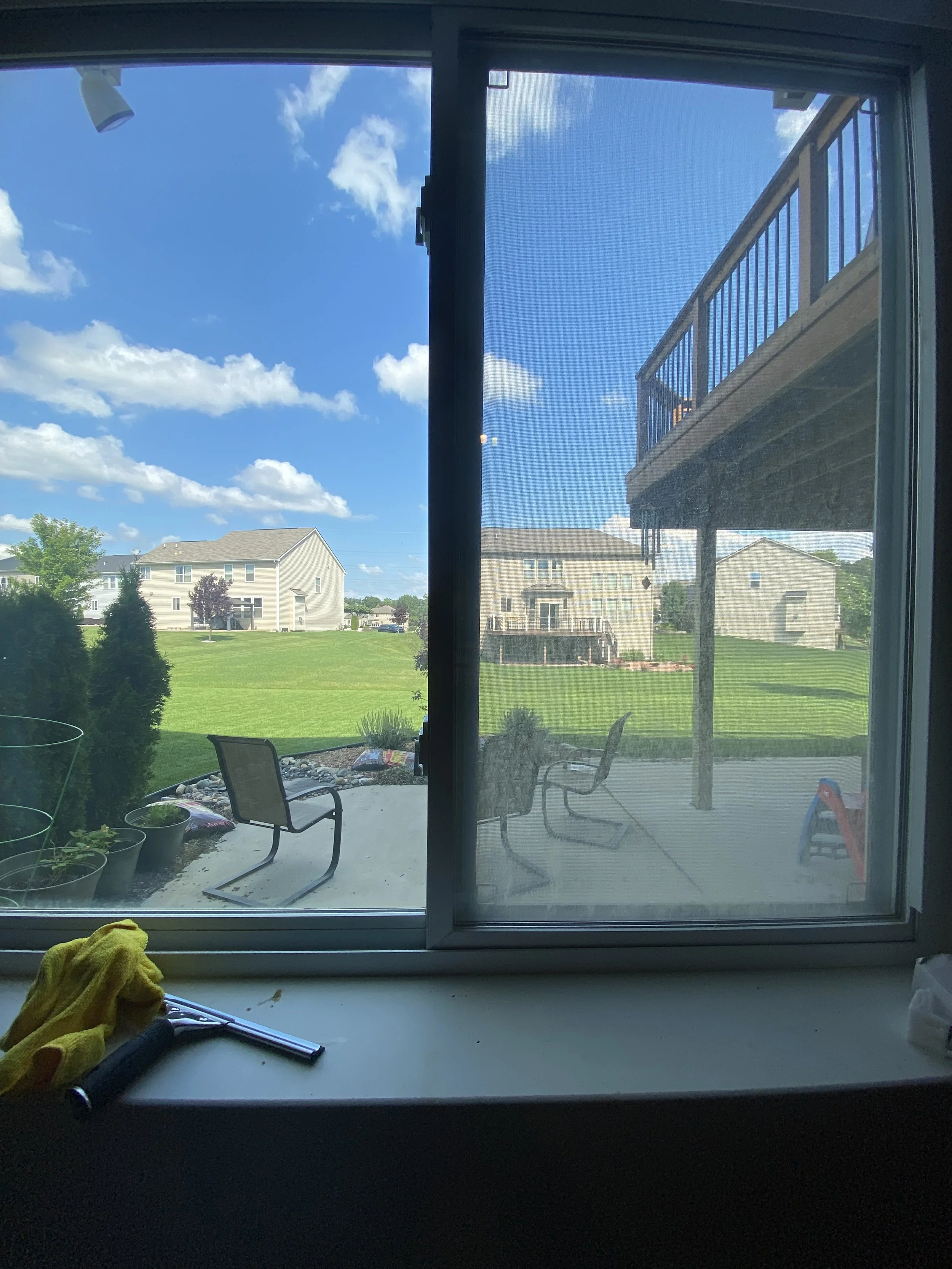 View through a window showing a sunny backyard with green grass, patio chairs, plants, and neighboring houses in the distance under a blue sky with scattered clouds.