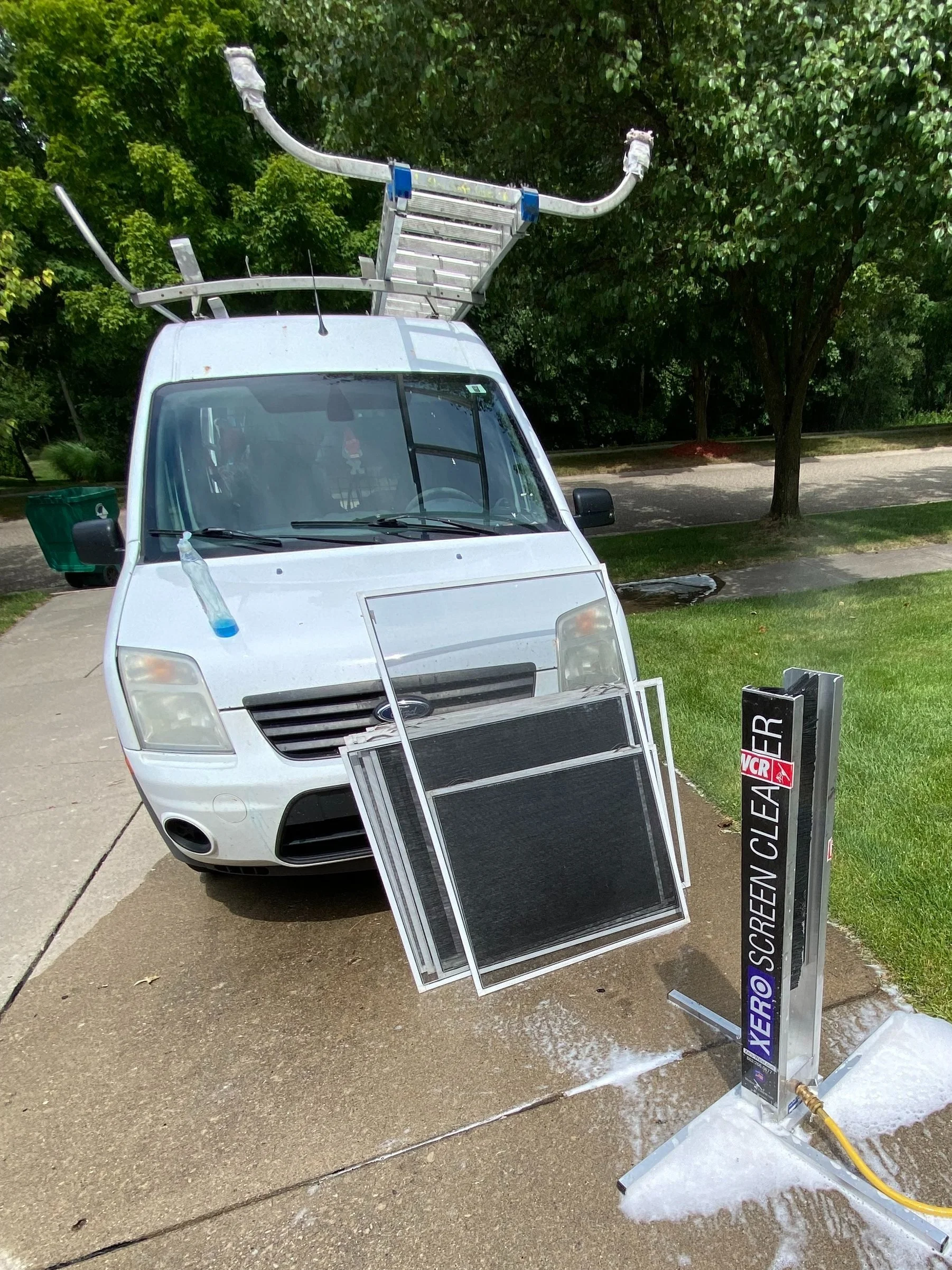 A white, small commercial van with ladders on the roof, soap and water on the driveway, and a screen cleaning machine labeled "Hero Screen Cleaner" in front of it, during daytime.