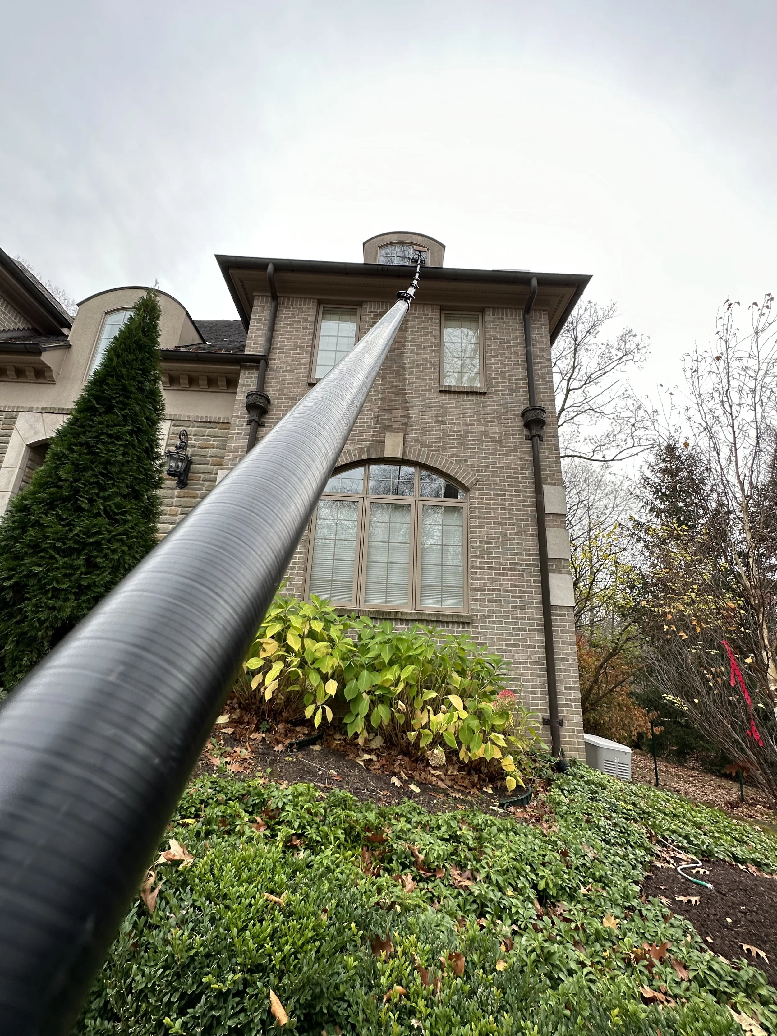 A view of a multi-story brick house from the front yard, with a tall black pole extending upward and leading to the house's roof. There are shrubs and trees in the garden area.