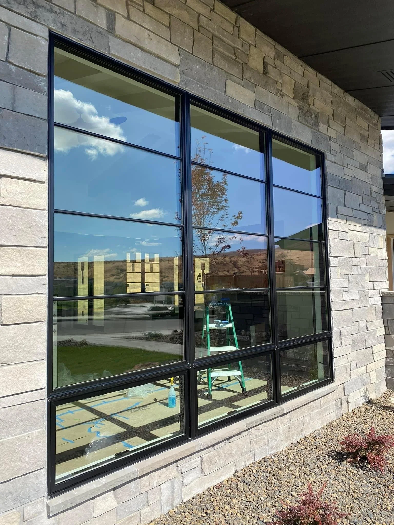 Large window with black framing showing reflections of sky, clouds, and trees, set in a stone wall with a gravel ground outside.
