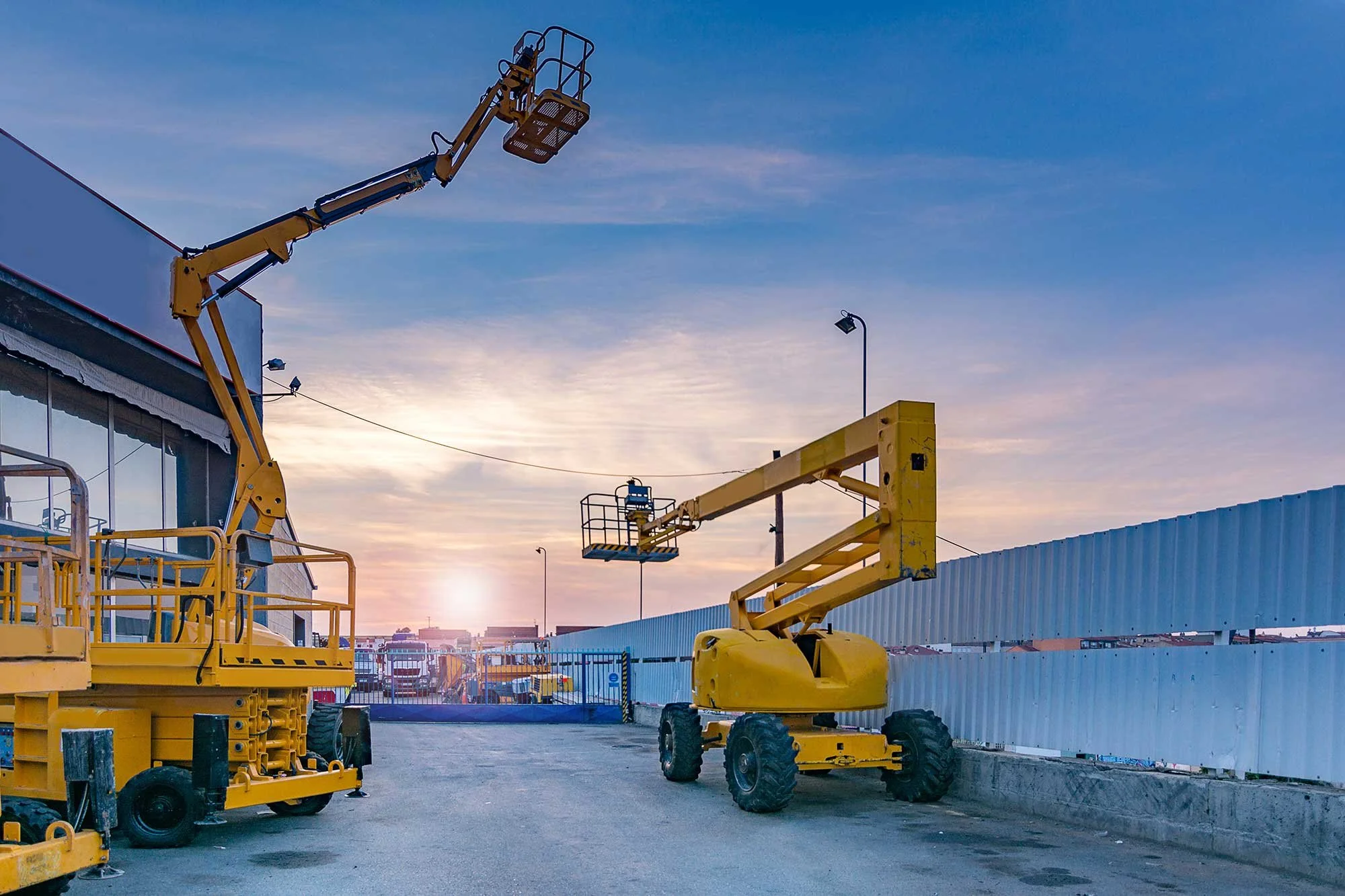 Construction site with yellow aerial lift equipment and a blue fence at sunset.