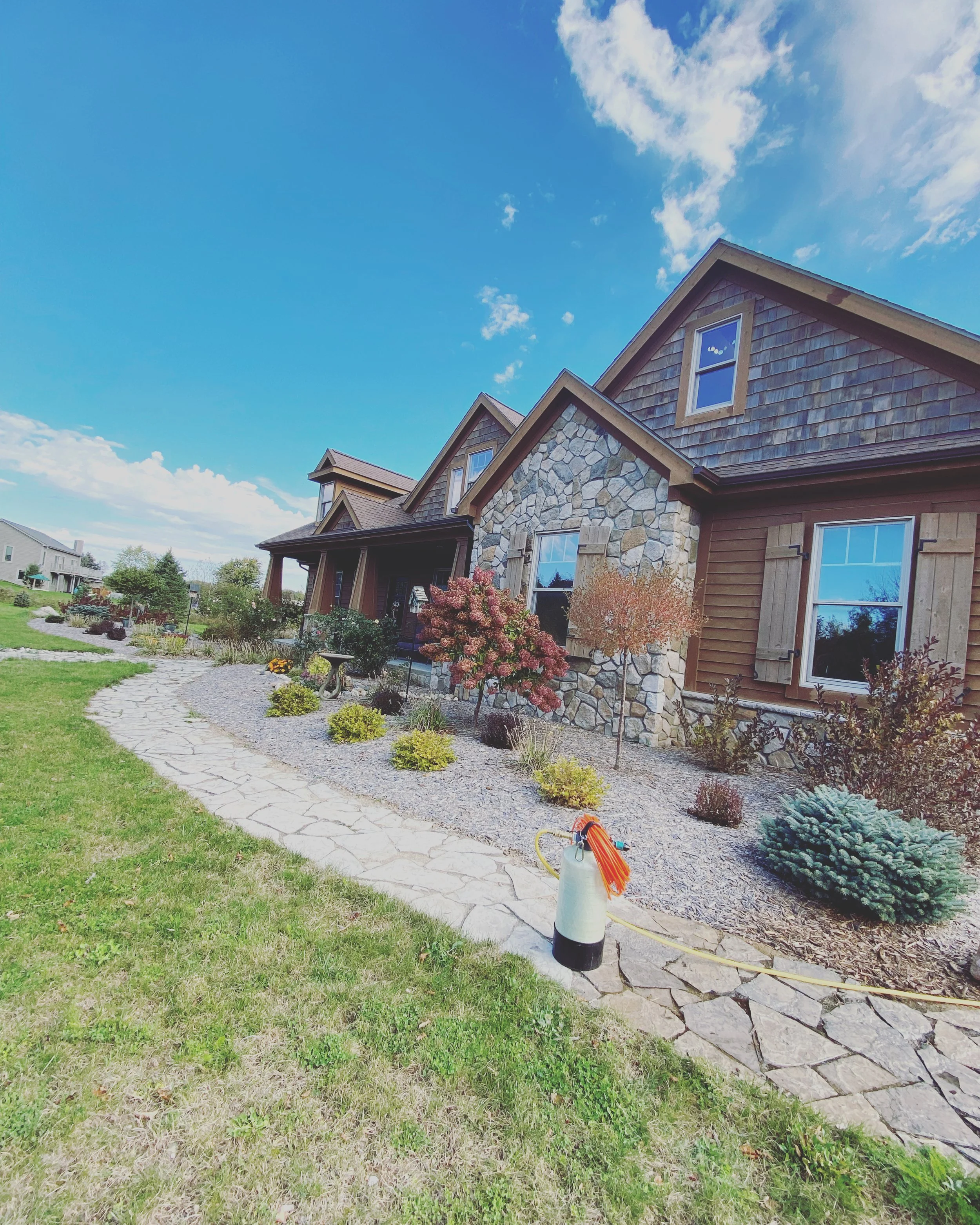 Front yard of a house with a stone and wooden exterior, landscaped with a stone pathway, small trees, bushes, and a lawn sprinkler on the grass.