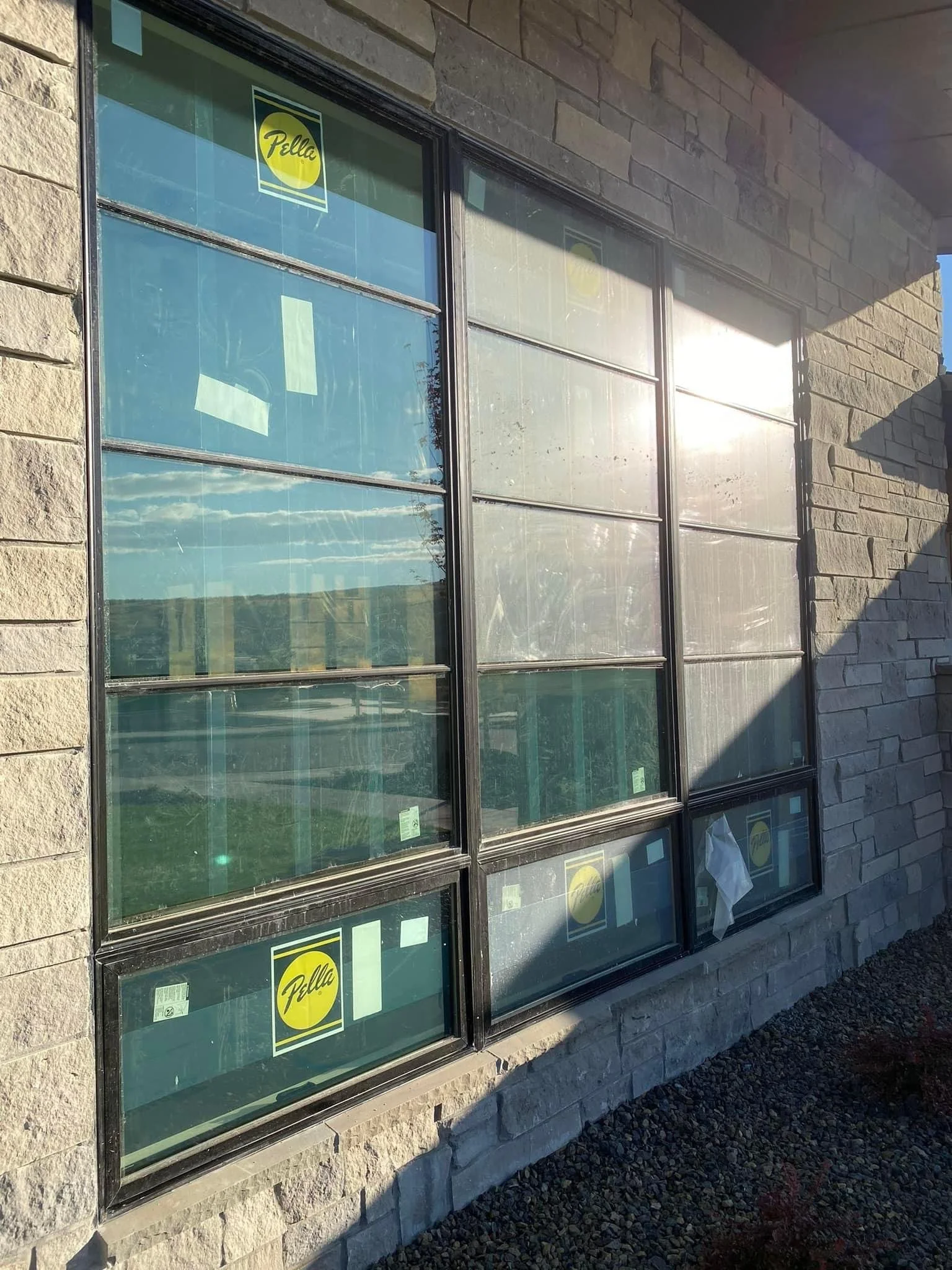 Storefront window with Pella logo stickers, reflecting the sky and landscape, set in a stone wall building exterior.