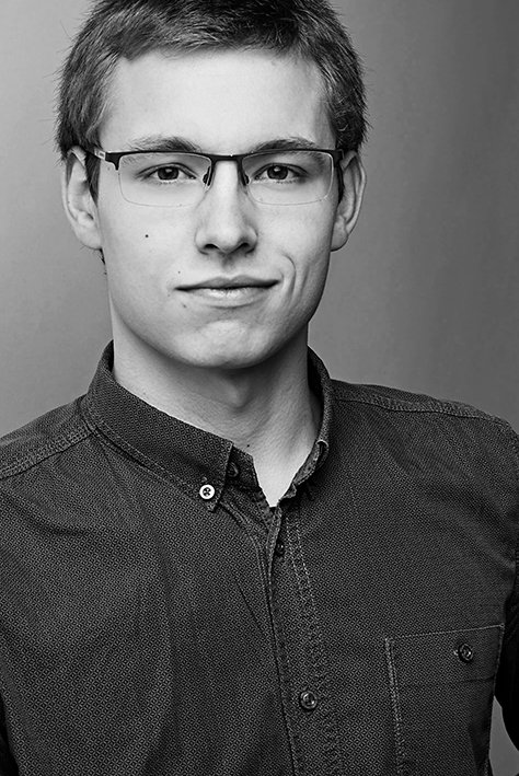 Schwarz-weiß Portrait eines jungen Mannes mit kurzen dunklen Haaren, Brille und einem dunklen Hemd, vor grauem Hintergrund.