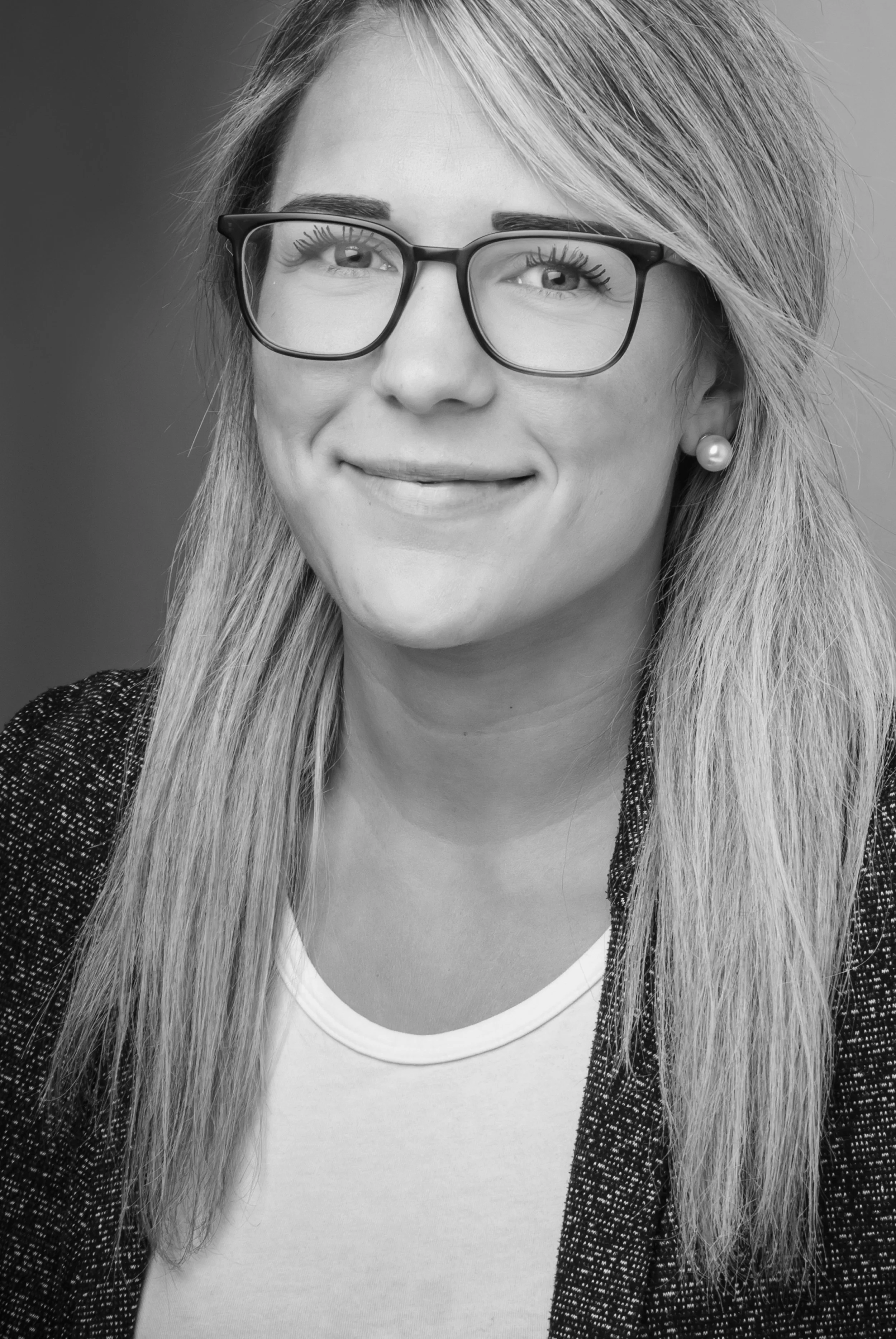 Schwarz-weiß Porträt einer lachenden Frau mit Brille, langen glatten Haaren, Perlenohrringen, weißem Top und dunklem Blazer vor hellem Hintergrund.