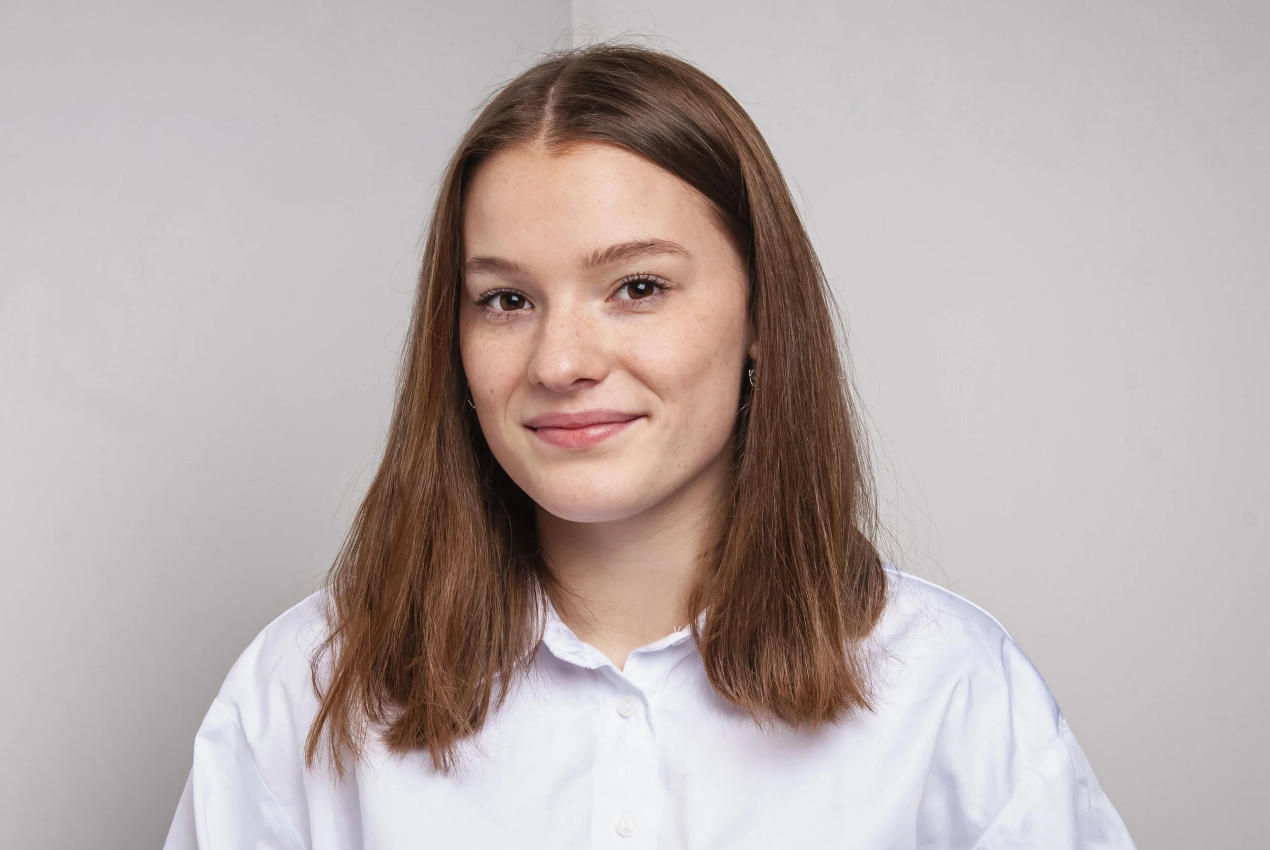 Porträt einer jungen weißen Frau mit braunen Haaren, die ein Weißes Hemd trägt, vor einem grauen Hintergrund.