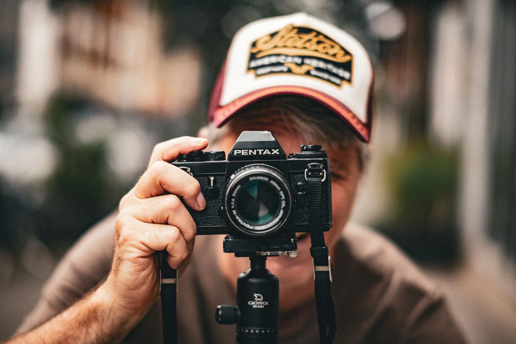 Jemand fotografiert mit einer Pentax-Kamera, während er auf einem Stativ steht, trägt einen Schirmmütze.
