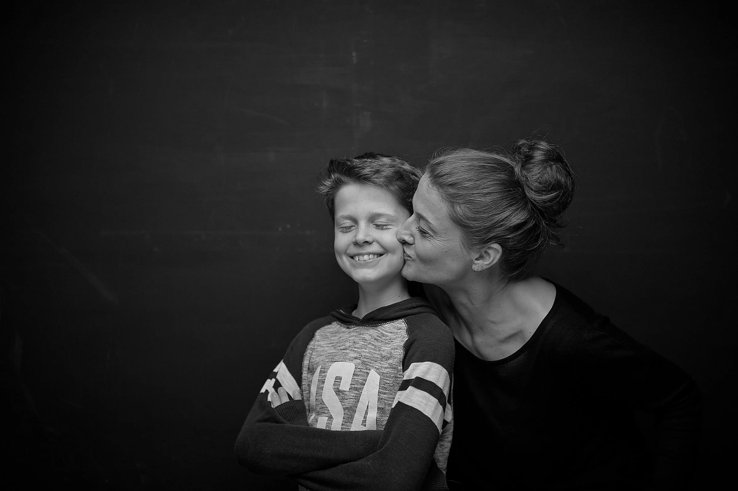 Emotionales Familienfoto einer Mutter küsst ihren lachenden Sohn auf die Wange, beide vor einem schwarzen Hintergrund im Fotostudio Hannover