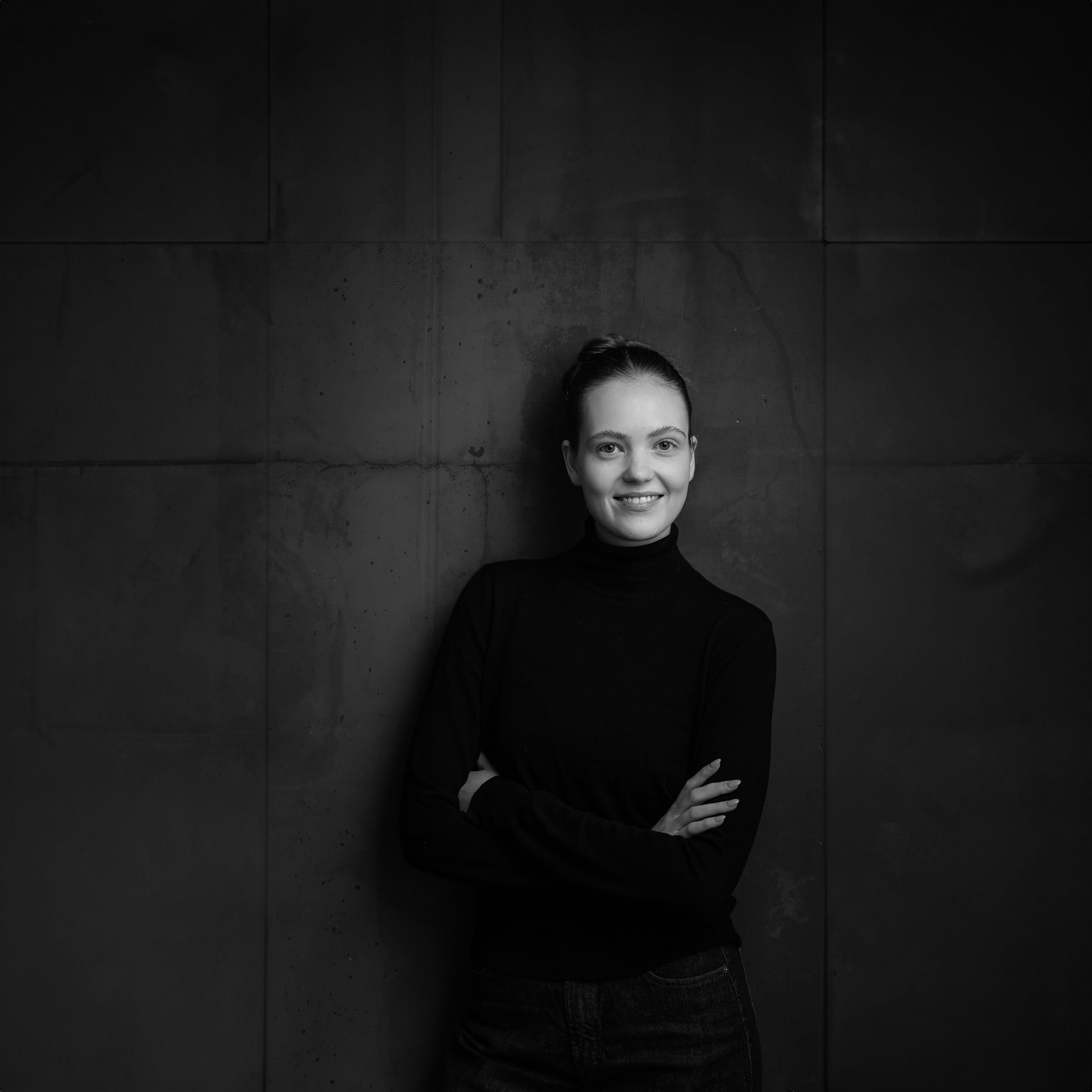 Junge Frau im schwarzen Pullover und Jeans steht vor einer dunklen Wand, lächelt und schaut in die Kamera. Professionelle Bewerbungsfotos im Fotostudio in Hannover