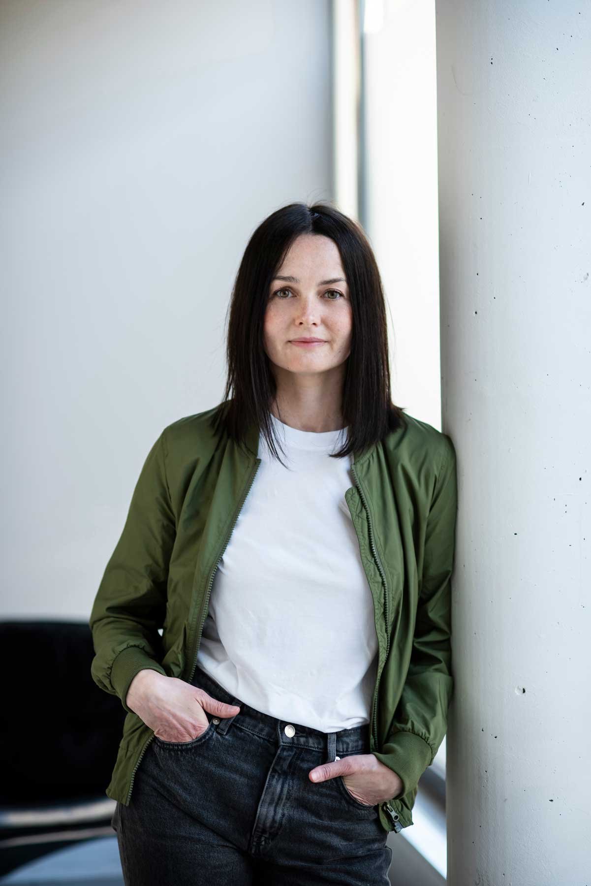 Professionelle Bewerbungsfotos im Fotostudio in Hannover. Eine Frau mit schwarzen Haaren, grüner Jacke und weißen T-Shirt lehnt an einer Säule in einem modernem Raum.