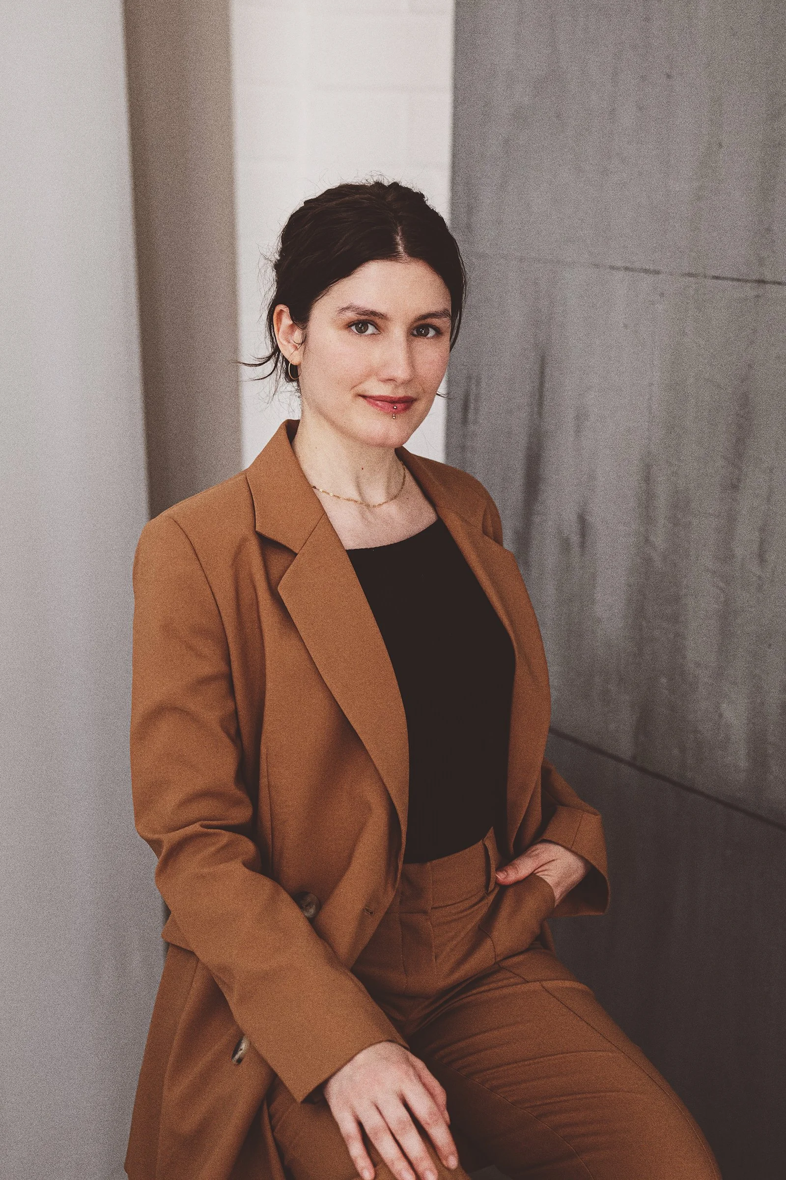 Junge Frau in brauner Anzug, sitzend vor grauem und weißem Hintergrund, lächelnd und mit kurzen dunklen Haaren, passende Accessoires, casual Businesslook.