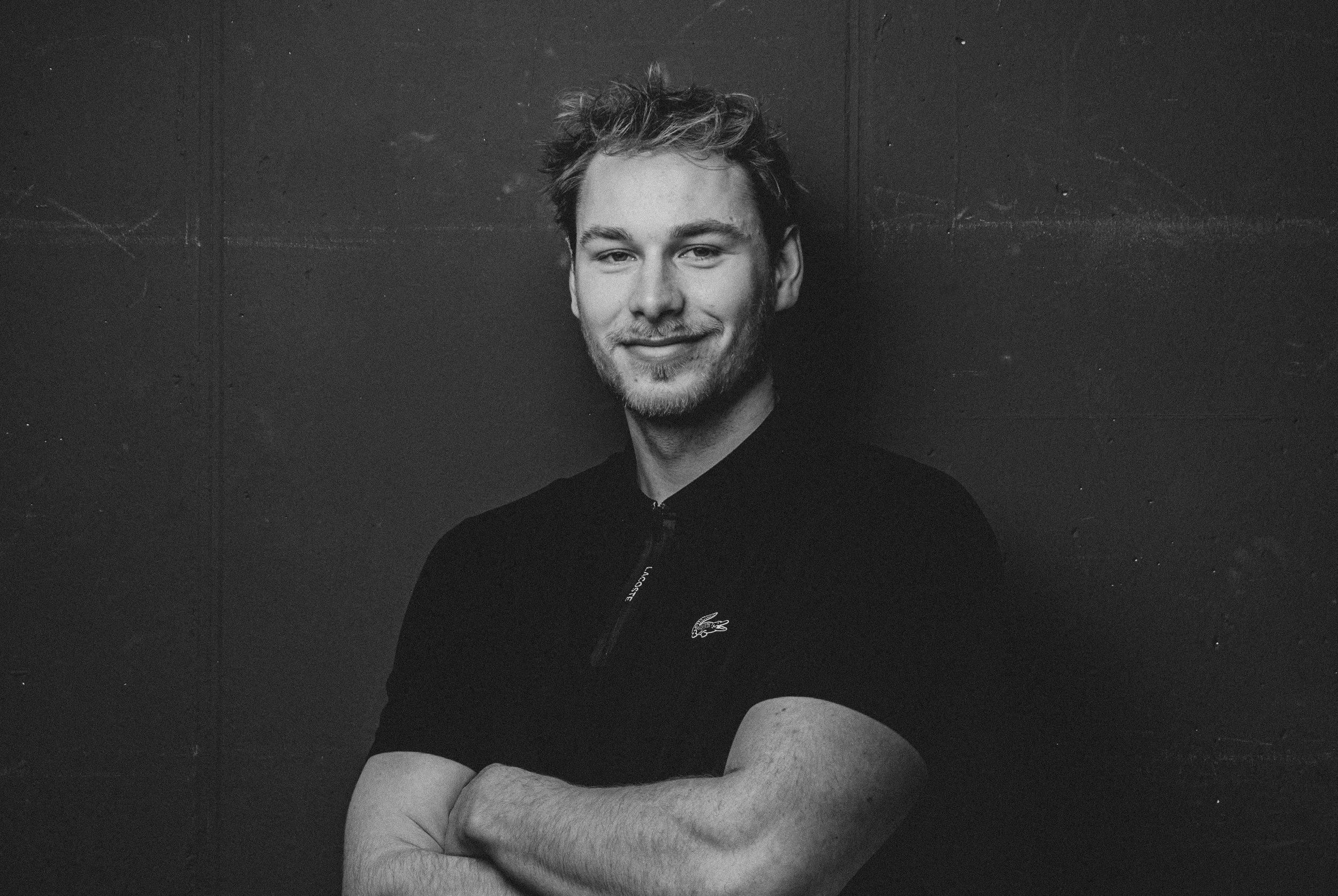 Schwarz-weiß-Fotografie eines lächelnden jungen Mannes mit kurze, unordentliche Haare und Rasierten Bart. Er trägt ein schwarzes Oberteil mit einem Lacoste-Logo und steht vor einer dunklen Wand. Professionelle Bewerbungsfotos im Fotostudio in Hannove