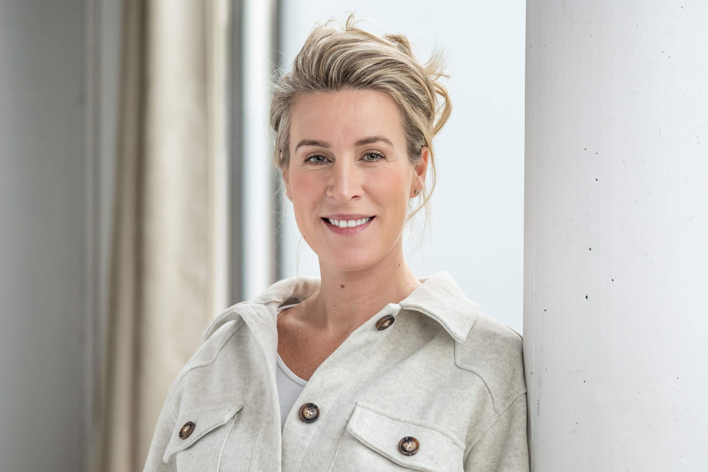 Nahaufnahme einer lachenden Frau mit kurzen blonden Haaren, die auf einem weißen Stein- oder Betonpfeiler steht, im Hintergrund sind helle Vorhänge. Professionelle Bewerbungsfotos im Fotostudio in Hannover