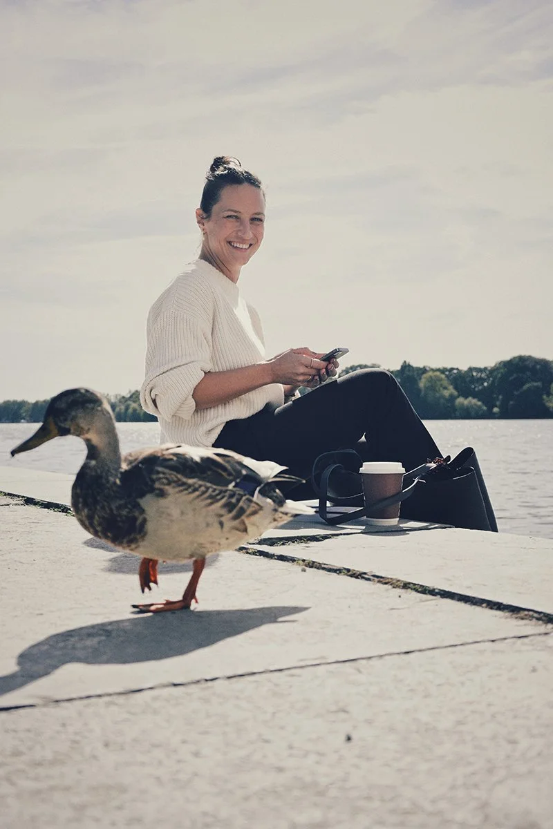 Eine lachende Frau sitzt am Flussufer mit einem Handy in der Hand, neben ihr ein Kaffee und Kopfhörer, im Vordergrund ein paarentenster Oberseite.