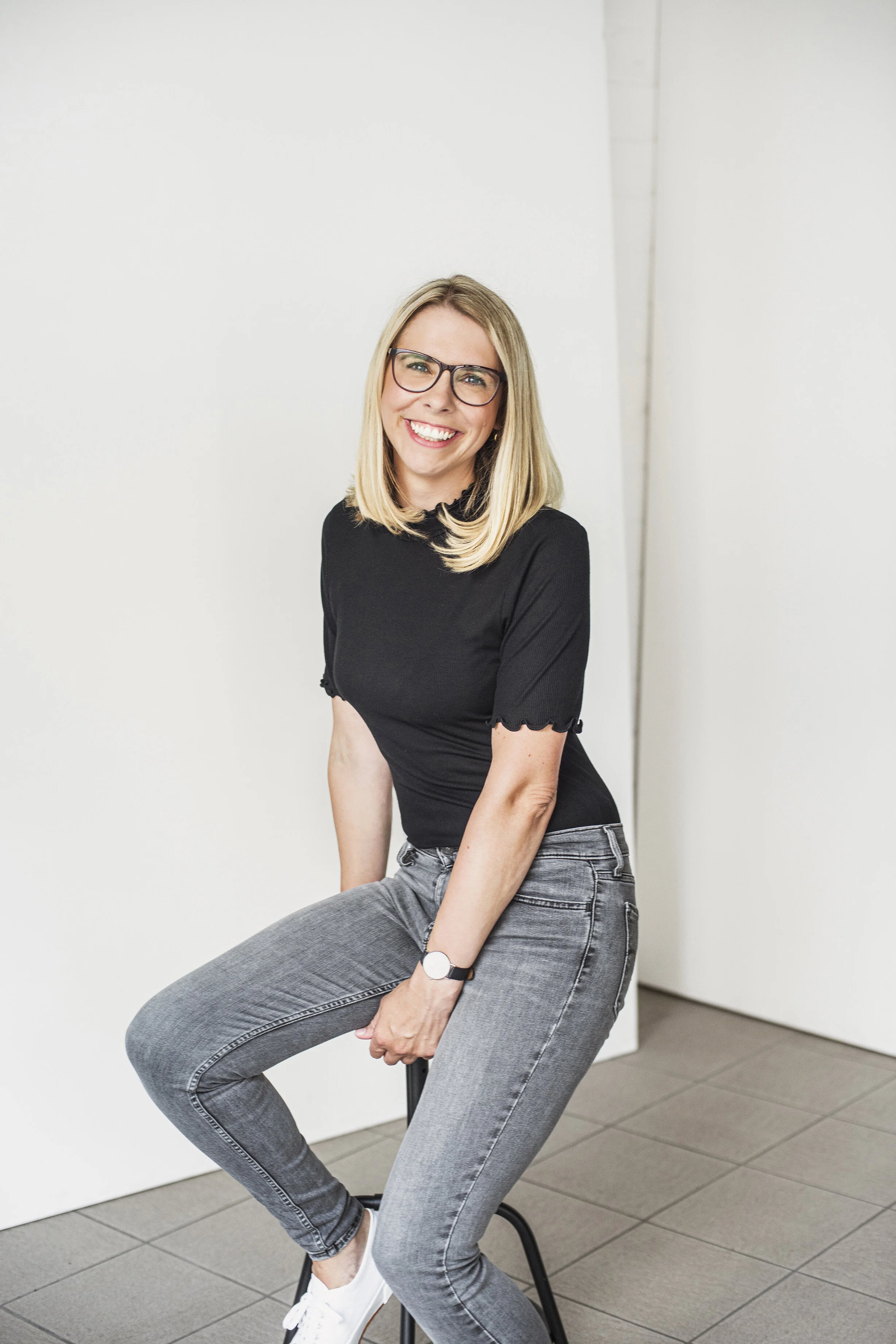 Eine Variante des selben professionellen Bewerbungsfotos im Fotostudio in Hannover von einer lächelnde Frau mit Brille die auf einem Stuhl vor weißer Wand in einem Raum sitzt.