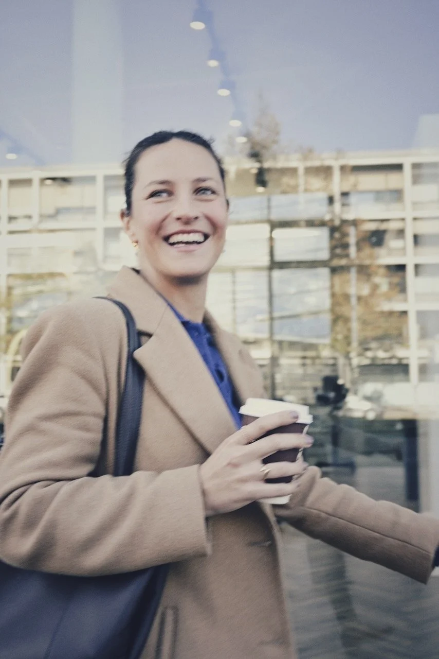 Eine lachende Frau mit braunem Mantel, die einen Becher Kaffee in der Hand hält, vor einem modernen Gebäudekomplex mit Glasfassade.