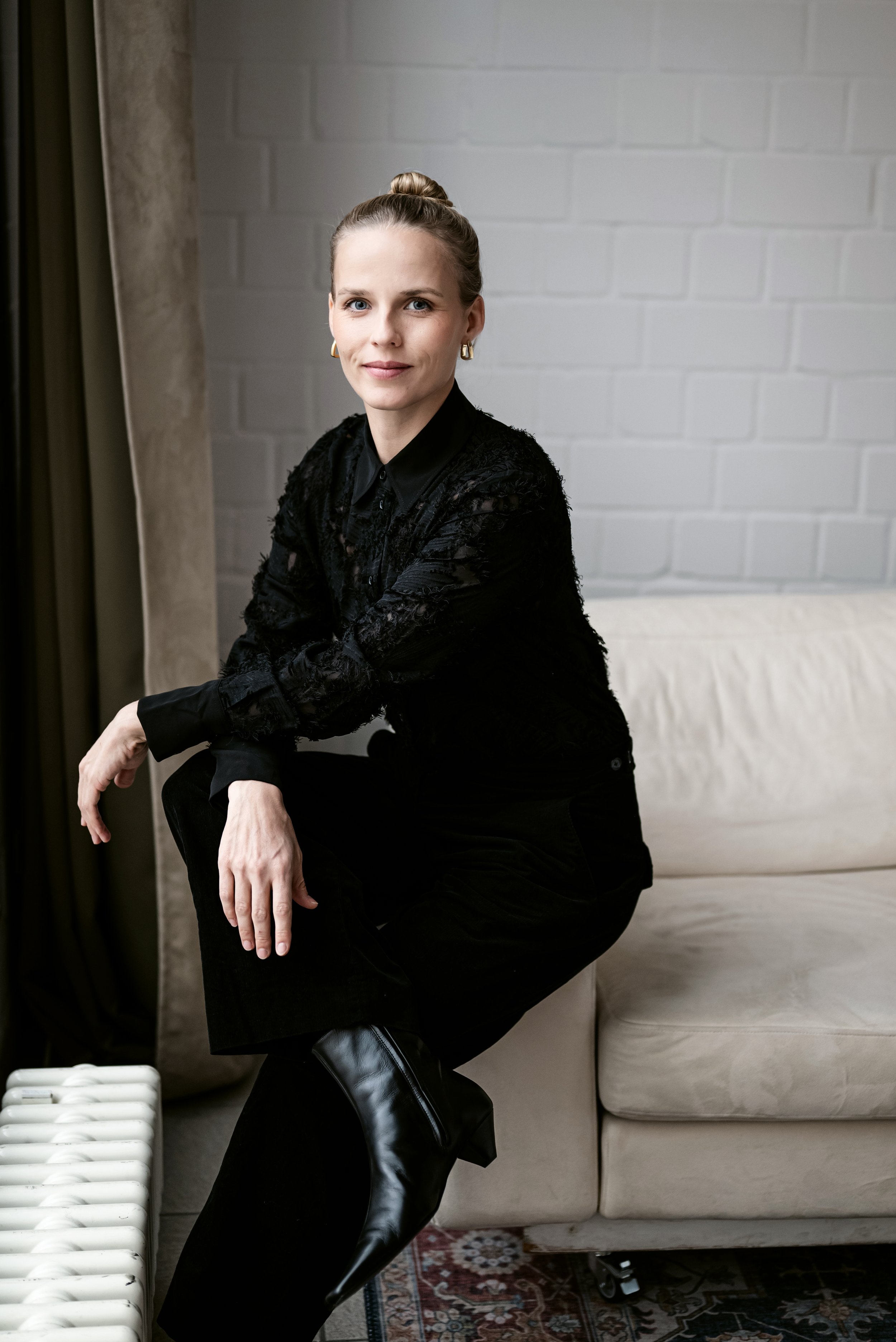 Eine junge Frau mit blondem Haar in einem schicken schwarzen Outfit sitzt auf einem Stuhl vor einer weißen Ziegelwand. Sie trägt Goldohrringe und lächelt subtil. Super schickes, modernes und professionelles Bewerbungsfotos im Fotostudio in Hannover