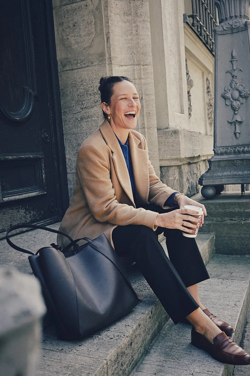 Eine lachende junge Frau sitzt auf den Stufen vor einem Gebäude mit Steinfassade und hält eine Tasse Kaffee in der Hand. Sie trägt eine beige Jacke, schwarze Hosen und braune Schuhe, neben ihr liegt eine große schwarze Tasche.