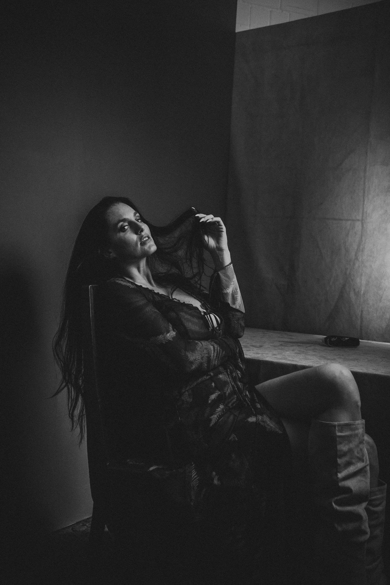 Schwarz-weiß-Bild einer jungen Frau mit langen Haaren, die auf einem Stuhl sitzt und in die Kamera schaut, während sie ihre Haare berührt. Im Hintergrund ein grauer Raum mit einer großen Leinwand oder Wand.