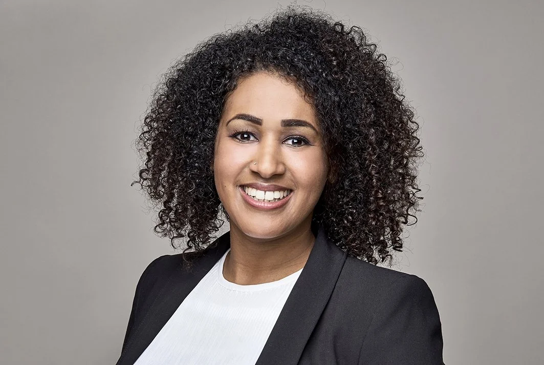 Porträt einer lachenden Frau mit lockigem dunkelbraunem Haar, dunkler Haut, in einem schwarzen Blazer und weißem Hemd vor grauem Hintergrund.