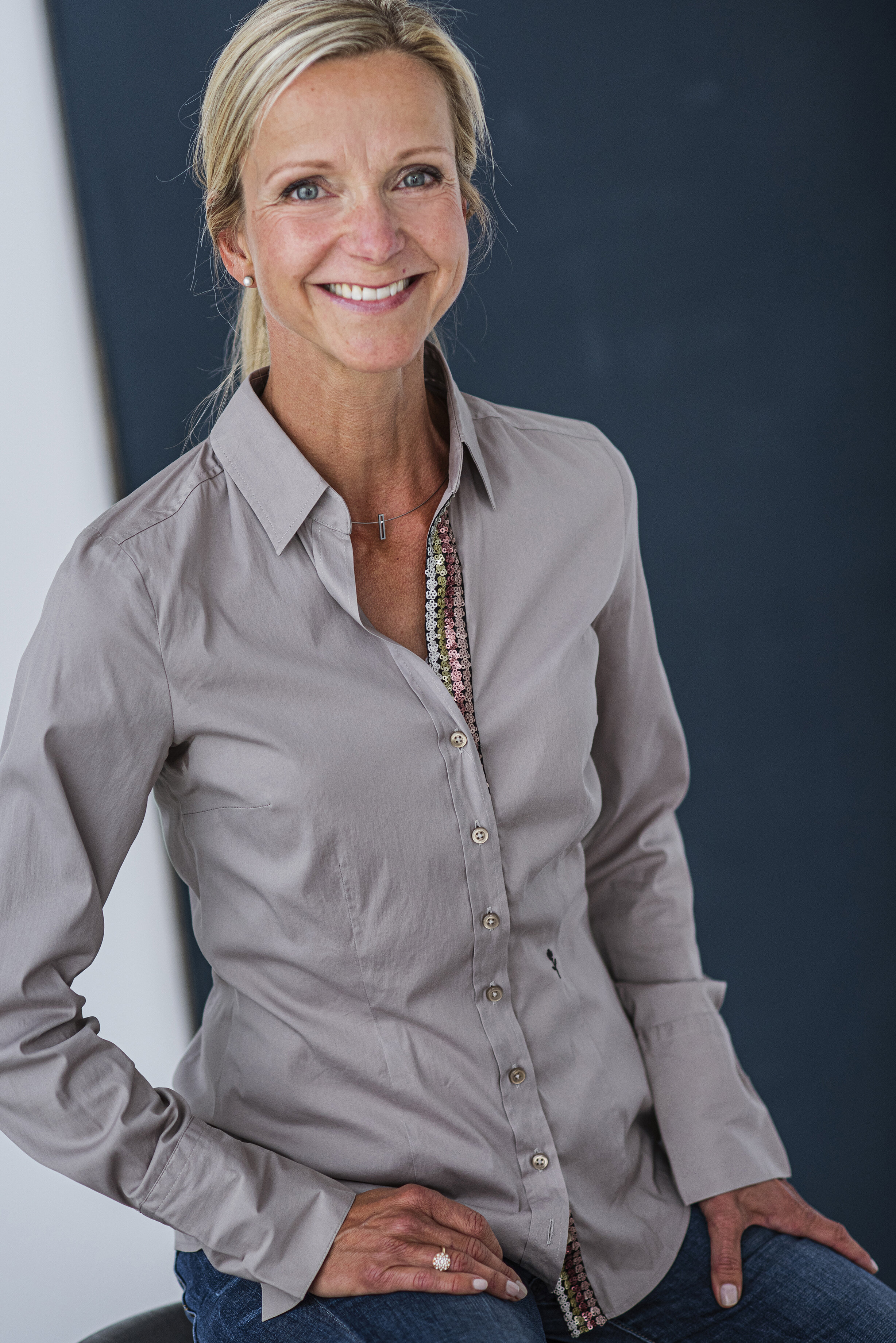 Die selbe lächelnde Frau mit blondem Haar, trägt eine graue Bluse und sitzt vor einem blauen Hintergrund. Professionelle Bewerbungsfotos im Fotostudio in Hannover